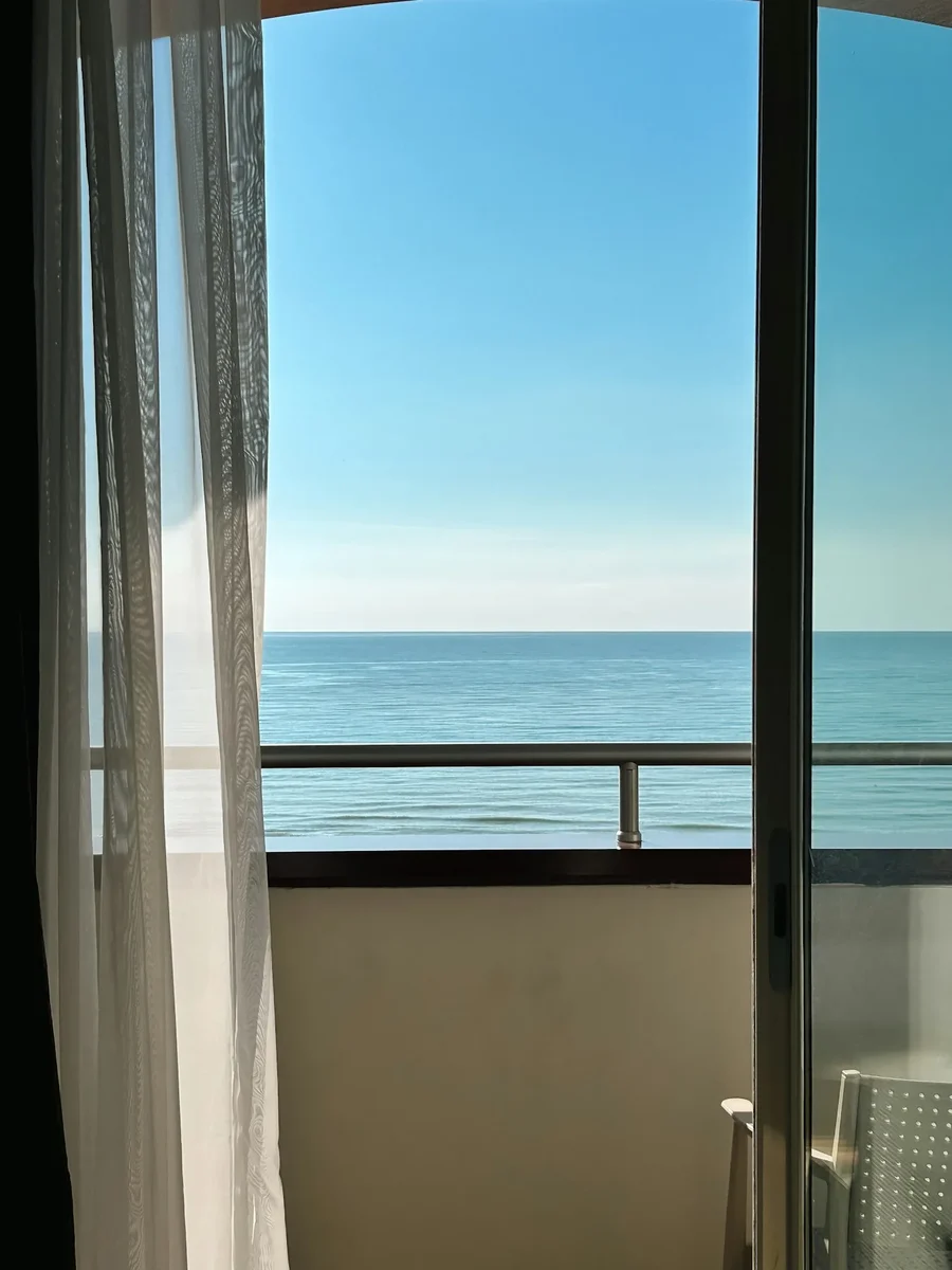 A sliding balcony door open to the sea with a sheer curtain drawn back