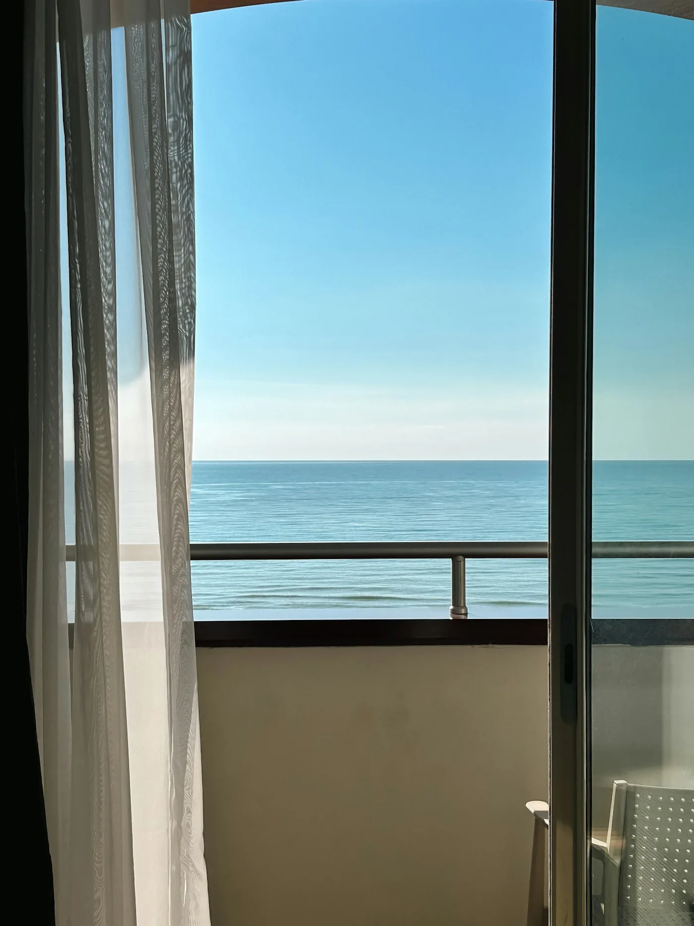 A sliding balcony door open to the sea with a sheer curtain drawn back