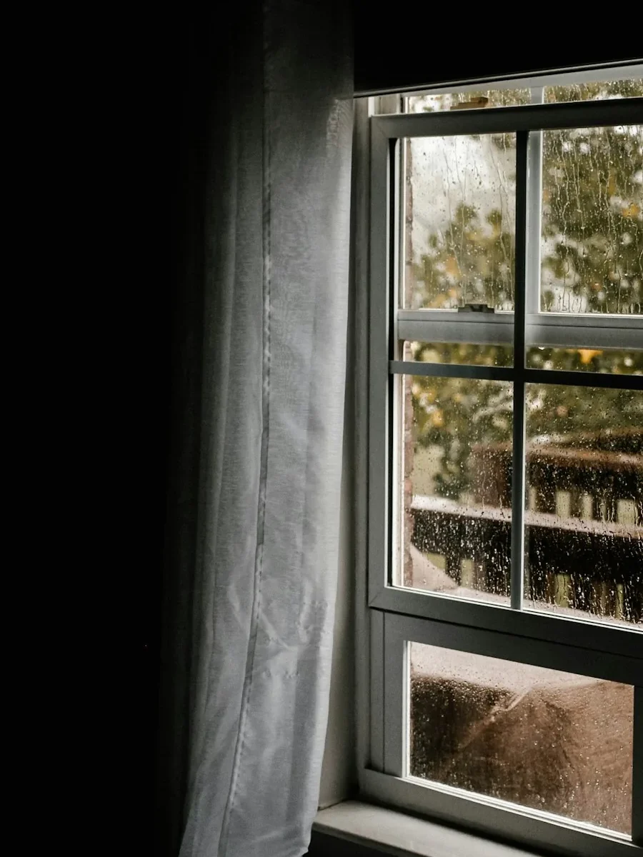 A dim window in a closed-up house, a sheer drawn across it and rain on the glass