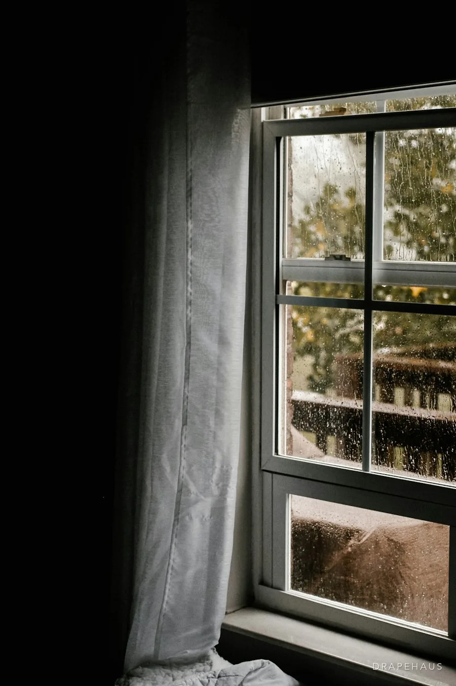 A dim window in a closed-up house, a sheer drawn across it and rain on the glass
