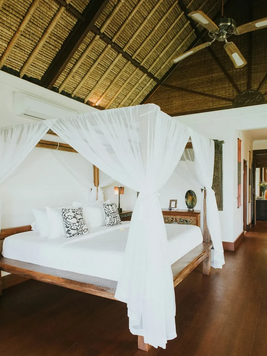 A tropical villa bedroom with a white-draped four-poster bed under a thatched roof and ceiling fan