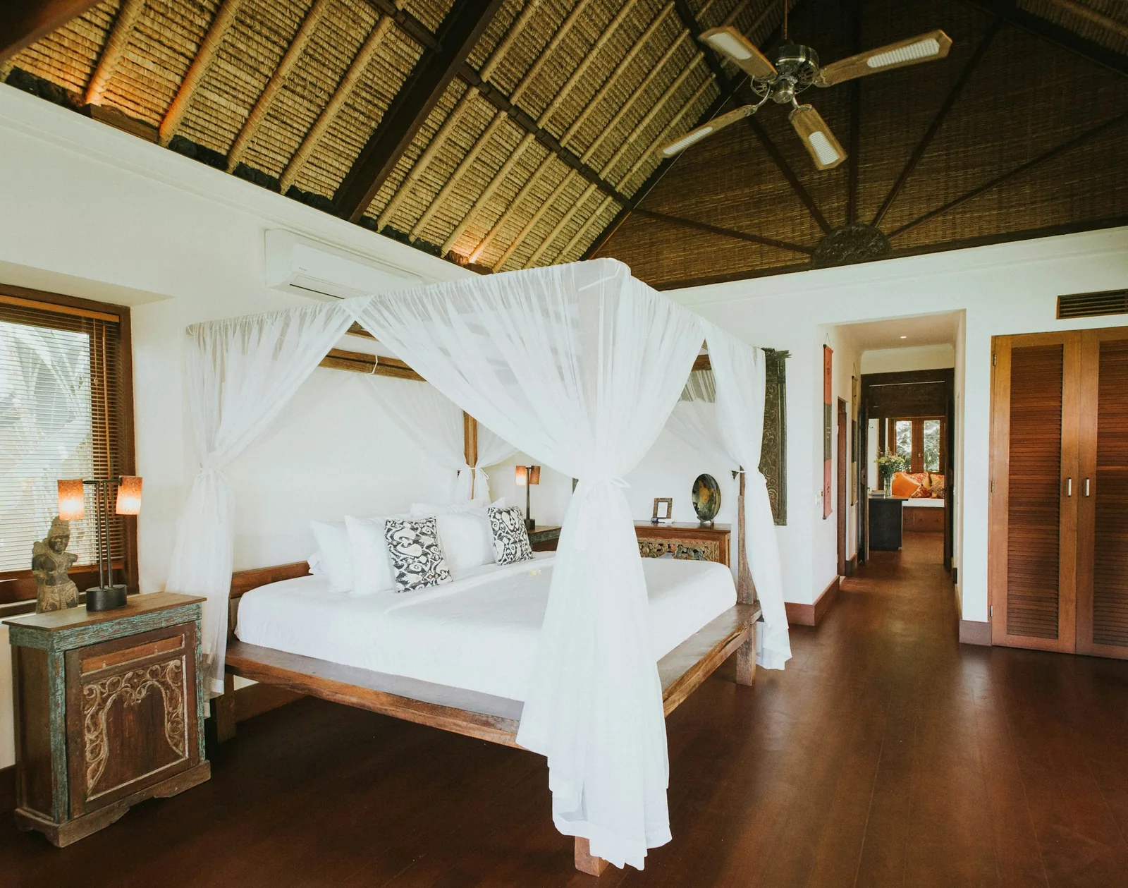 A tropical villa bedroom with a white-draped four-poster bed under a thatched roof and ceiling fan