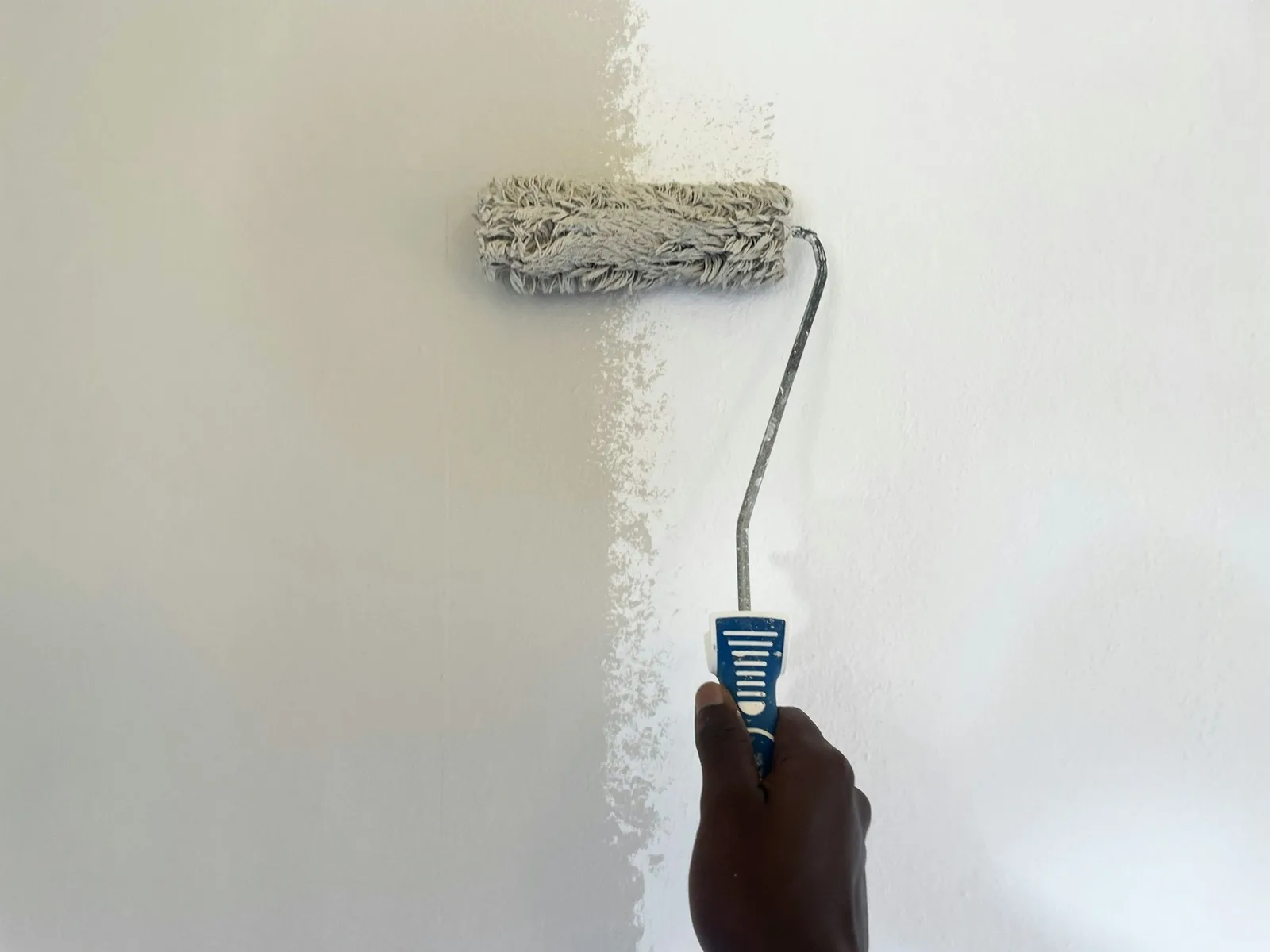 A hand rolling fresh white paint over a pale wall with a long-pile paint roller