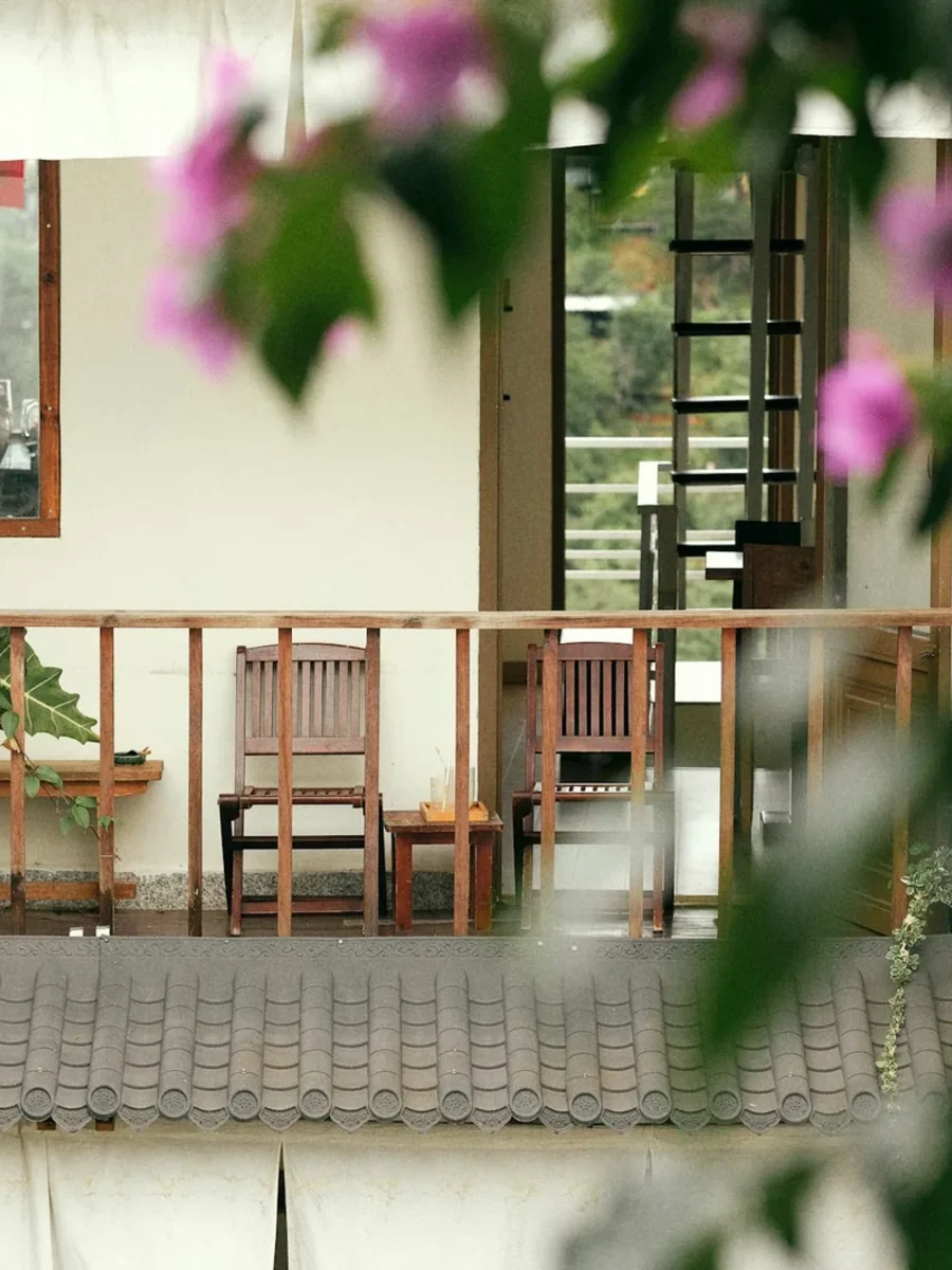 Small balcony with wooden railing, two chairs and potted plants, framed by pink bougainvillea