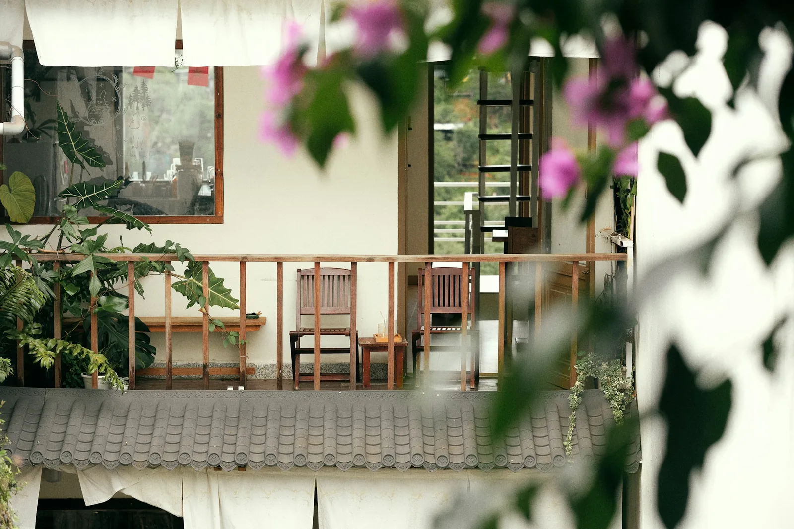 Small balcony with wooden railing, two chairs and potted plants, framed by pink bougainvillea