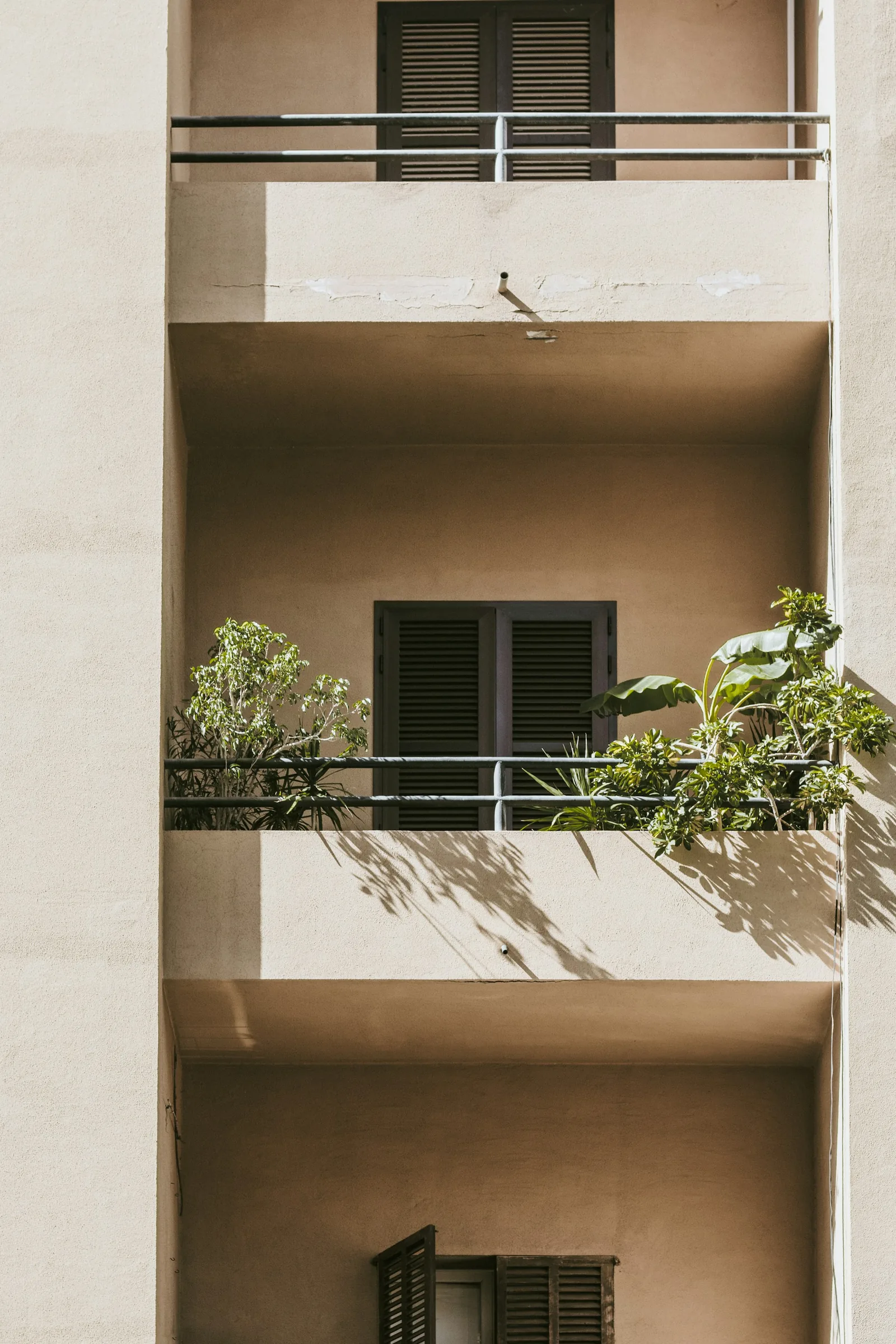 Stacked apartment balconies in warm beige render with dark louvred shutters and potted plants