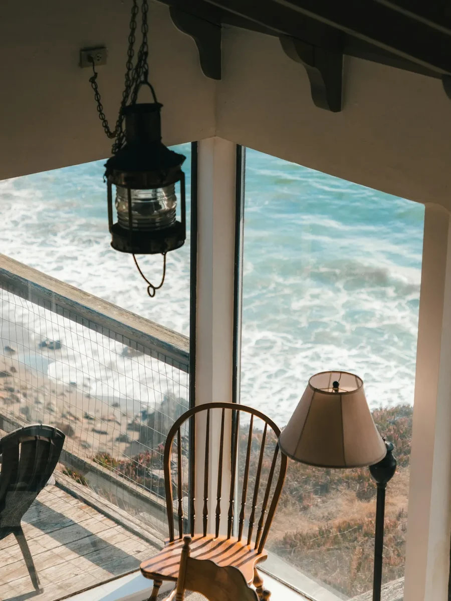 A corner window with a lantern and wooden chairs looking down onto waves breaking on a rocky shore