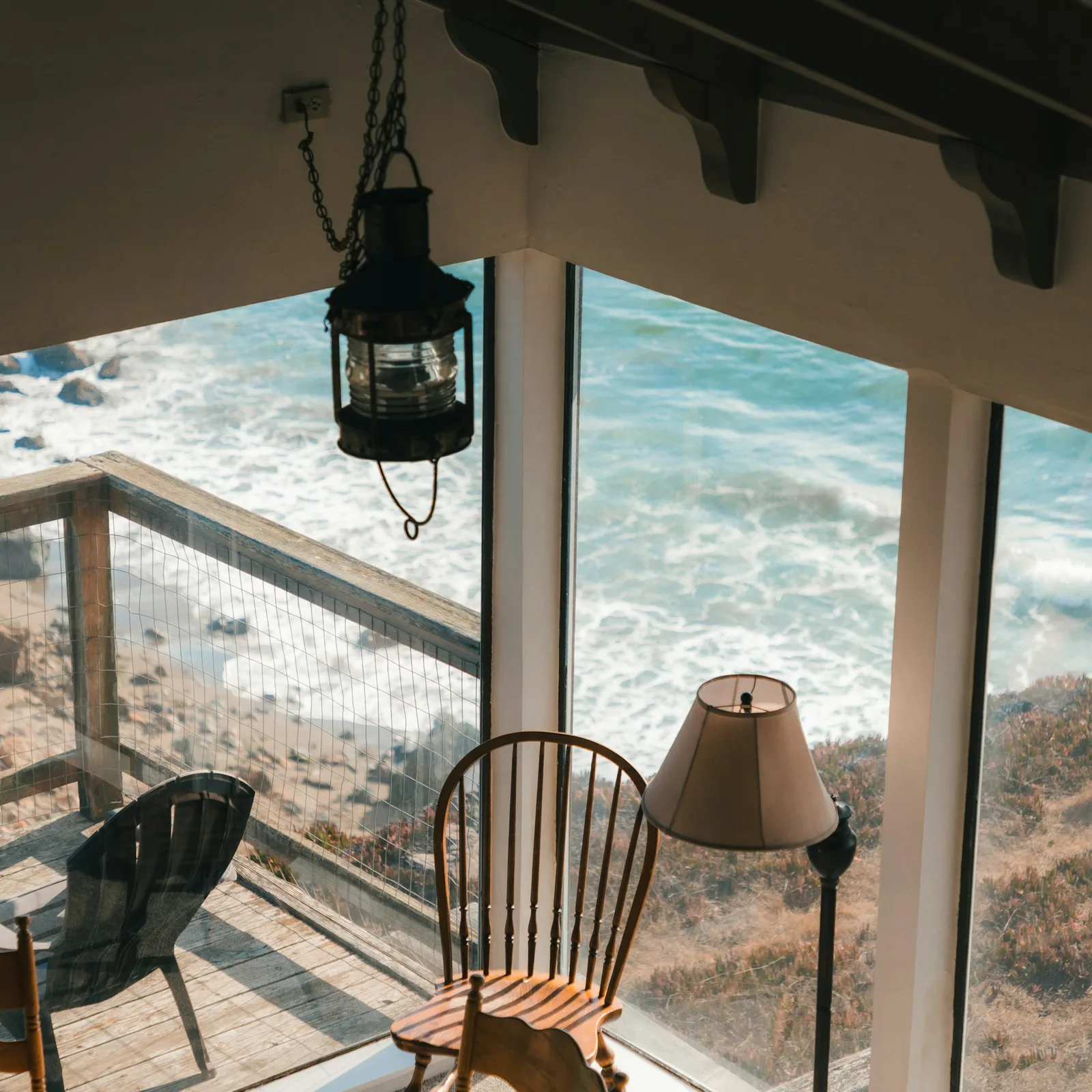 A corner window with a lantern and wooden chairs looking down onto waves breaking on a rocky shore
