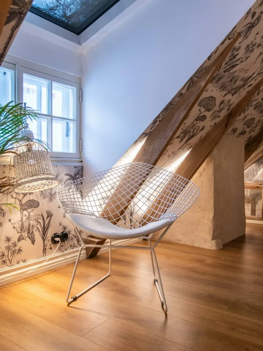 Attic sitting nook papered in a grey botanical wallpaper, with exposed beams and a wire chair