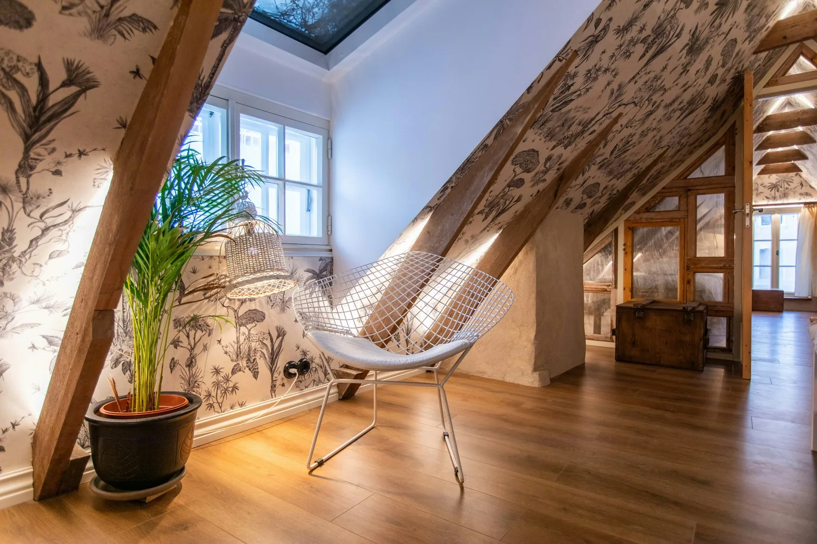 Attic sitting nook papered in a grey botanical wallpaper, with exposed beams and a wire chair