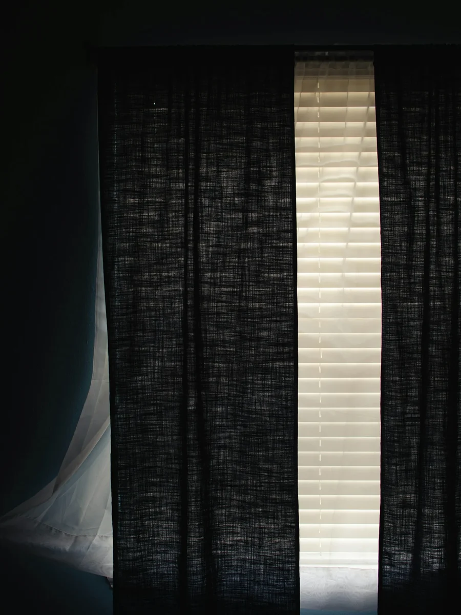 A dark linen curtain drawn across a window in a dim room, a thin strip of daylight at its edge