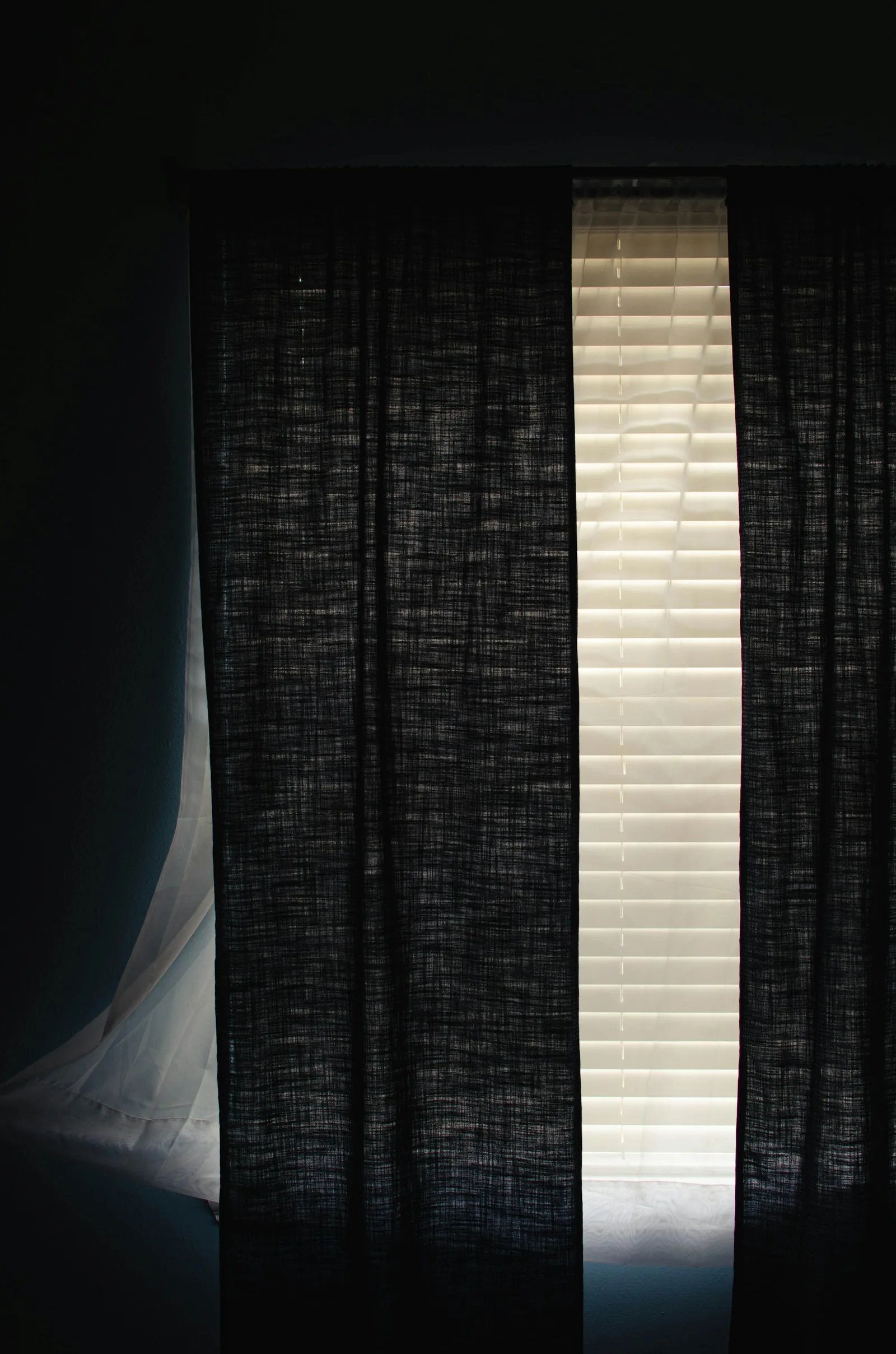 A dark linen curtain drawn across a window in a dim room, a thin strip of daylight at its edge