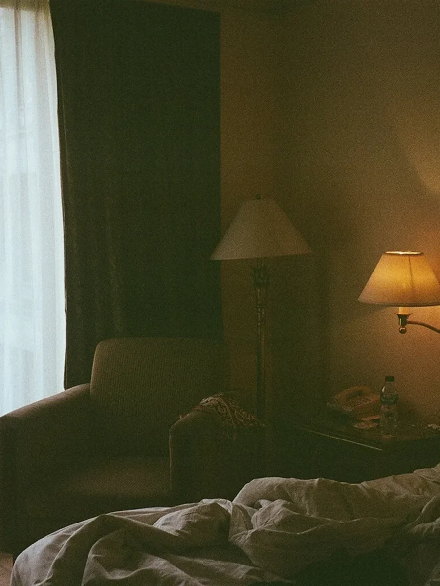 A dim bedroom with dark side curtains drawn back from sheers, an armchair and lamps lit beside the bed