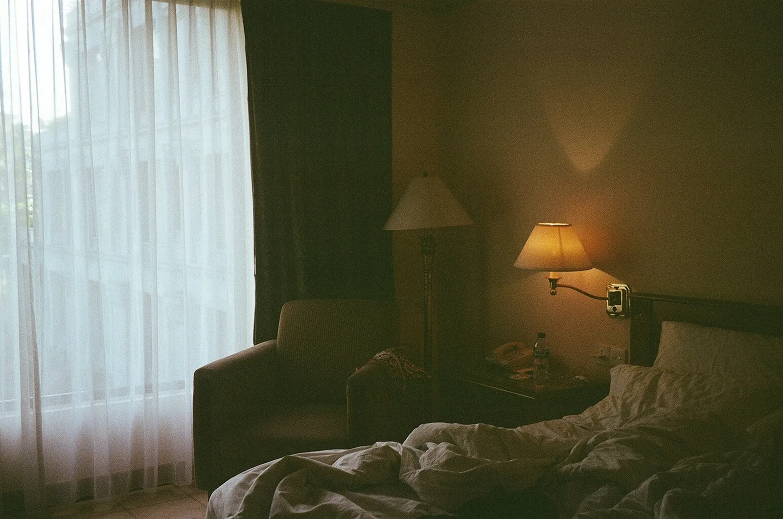 A dim bedroom with dark side curtains drawn back from sheers, an armchair and lamps lit beside the bed