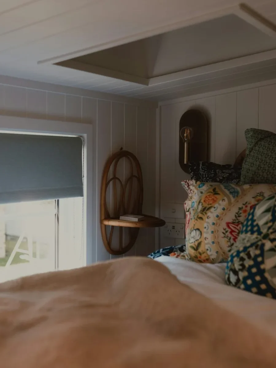 A grey roller blind half-lowered over a bedroom window beside a bed piled with patterned cushions