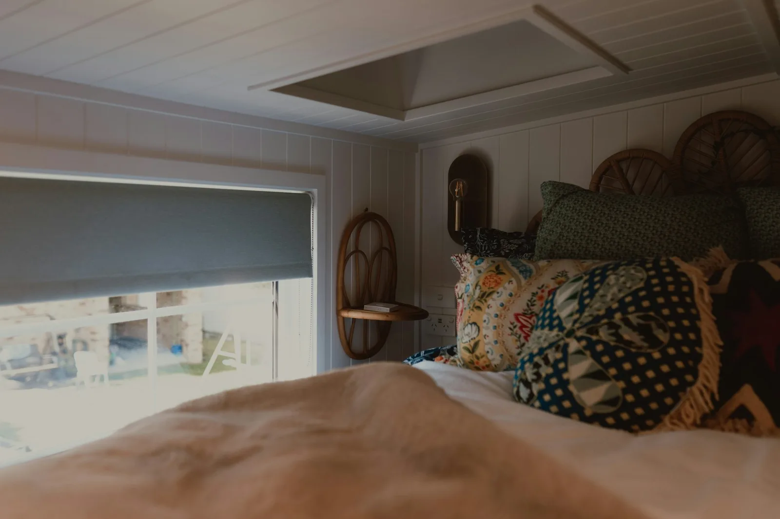 A grey roller blind half-lowered over a bedroom window beside a bed piled with patterned cushions