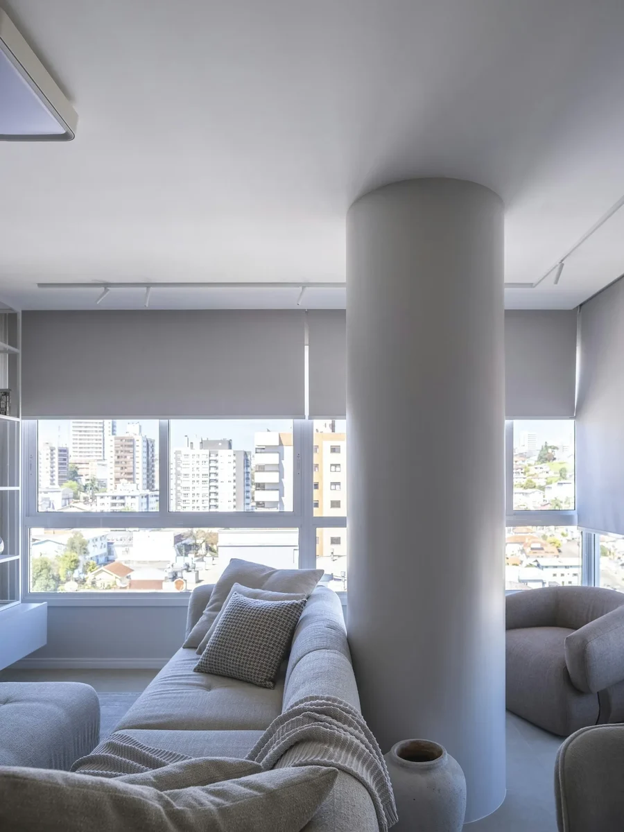 Grey roller shades half-lowered over wide windows in a pale living room with a sofa and round column