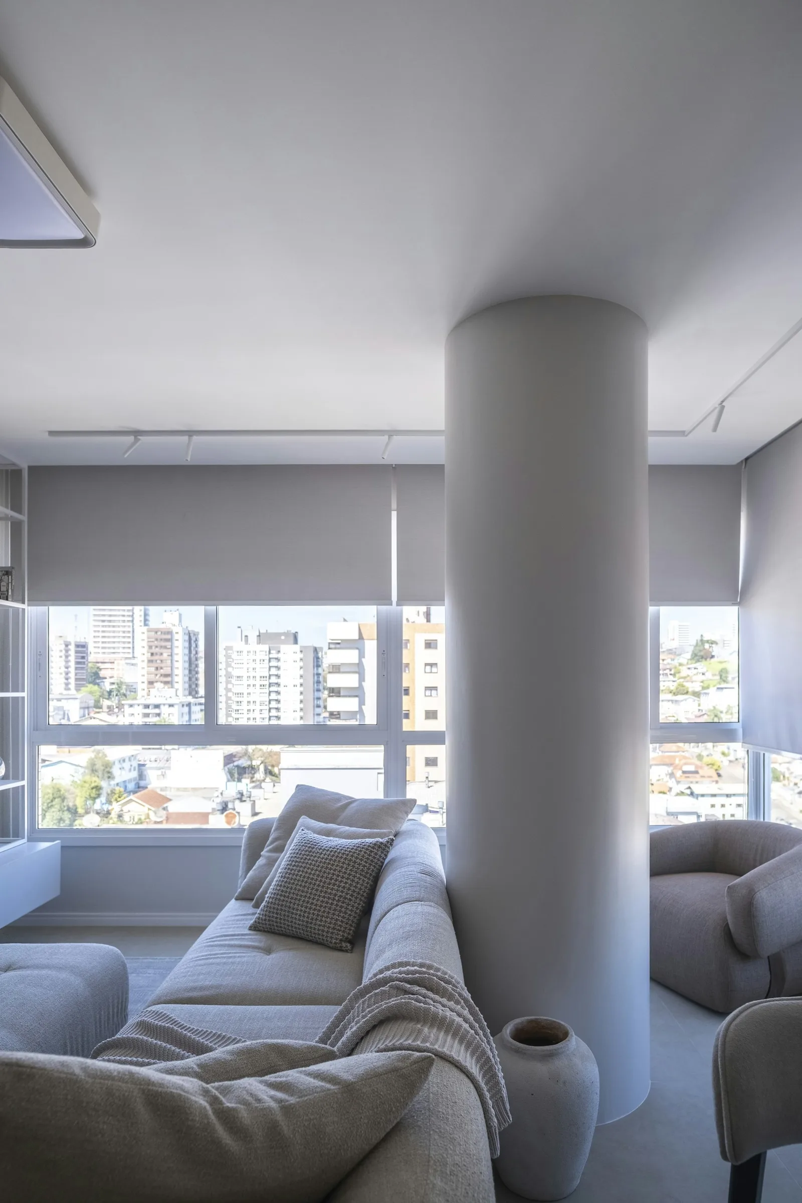 Grey roller shades half-lowered over wide windows in a pale living room with a sofa and round column