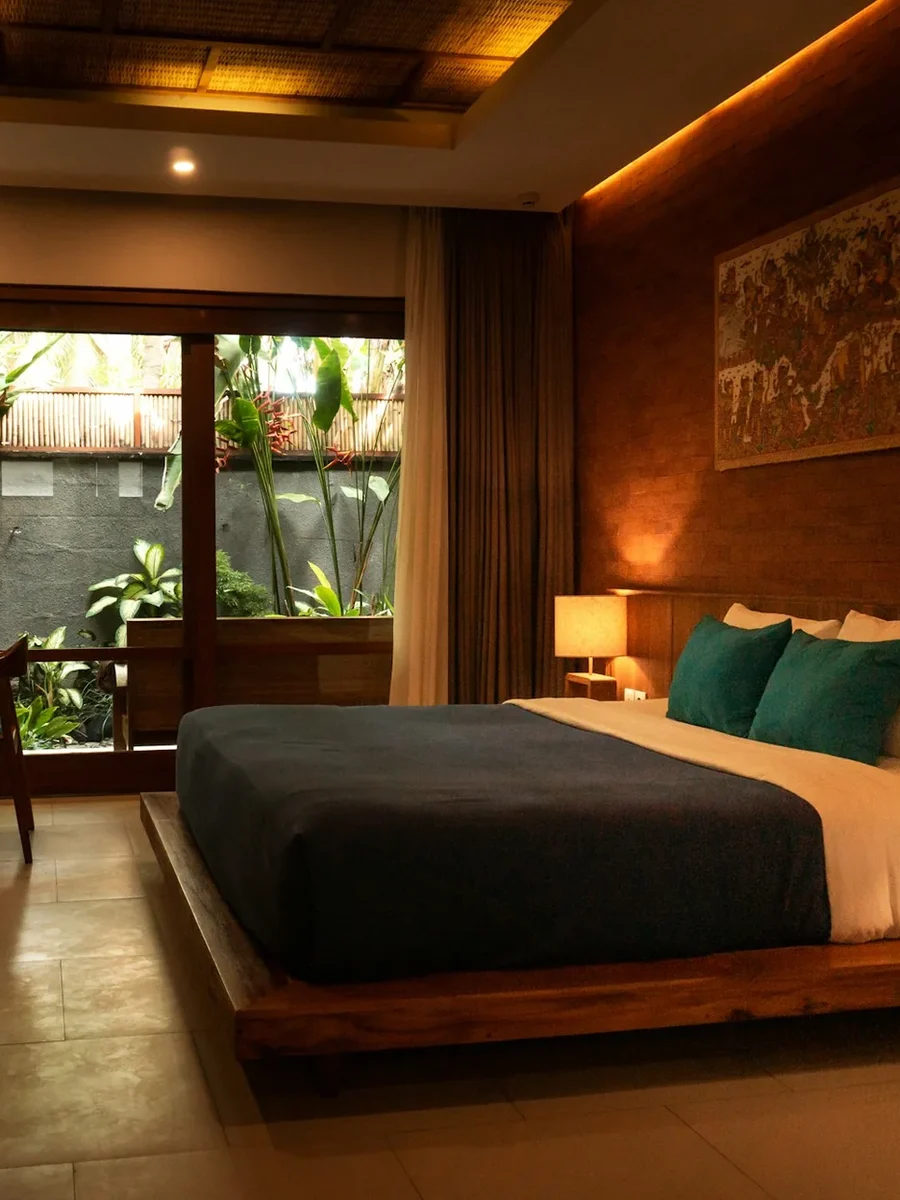 A warm hotel bedroom with timber panelling, teal cushions and glass doors onto a private tropical garden