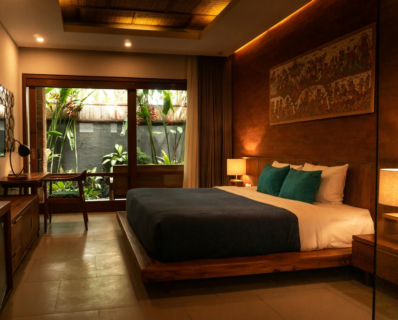 A warm hotel bedroom with timber panelling, teal cushions and glass doors onto a private tropical garden