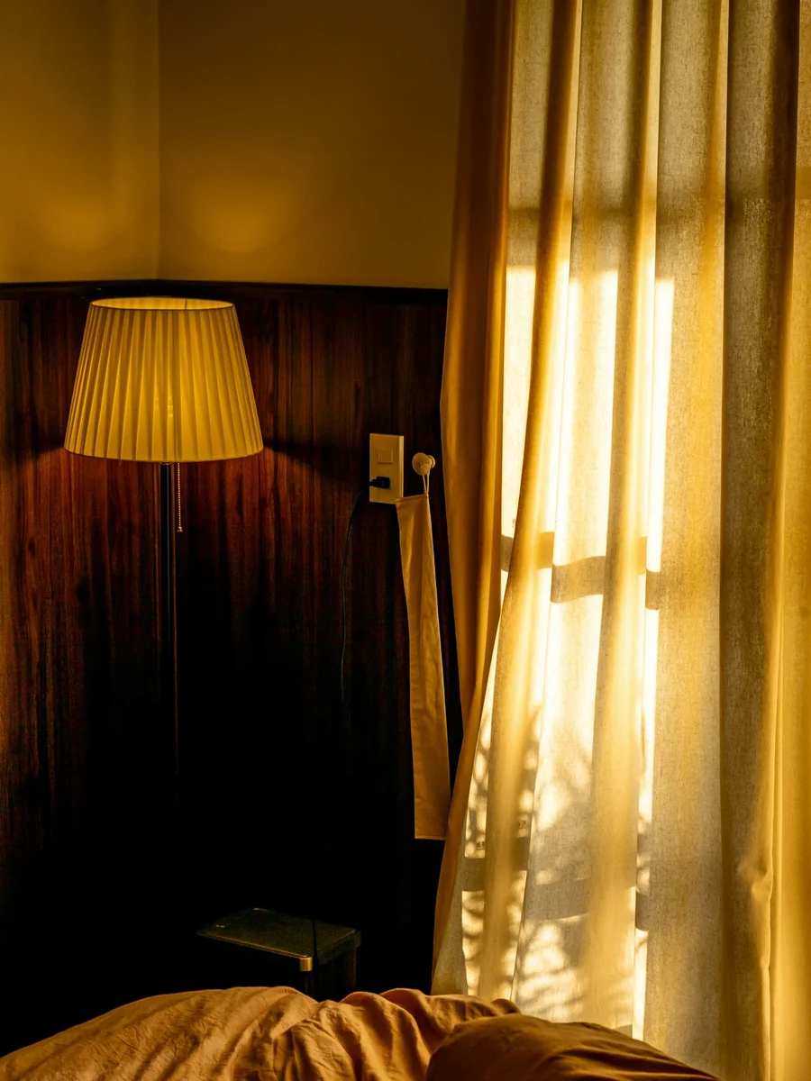 Golden-yellow curtains glowing in low sun beside a pleated lamp and a rumpled bed