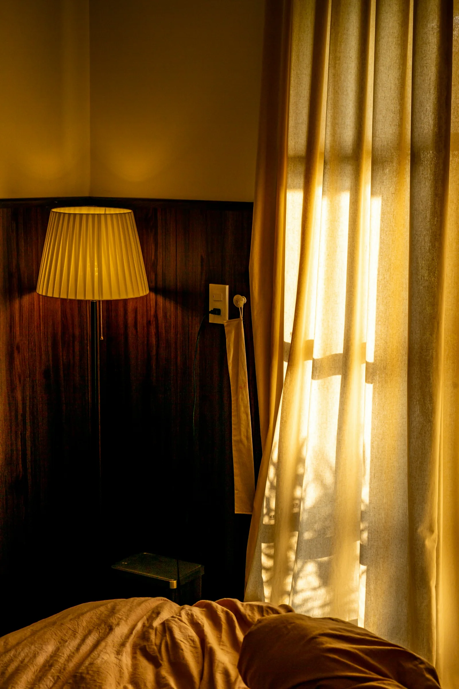 Golden-yellow curtains glowing in low sun beside a pleated lamp and a rumpled bed