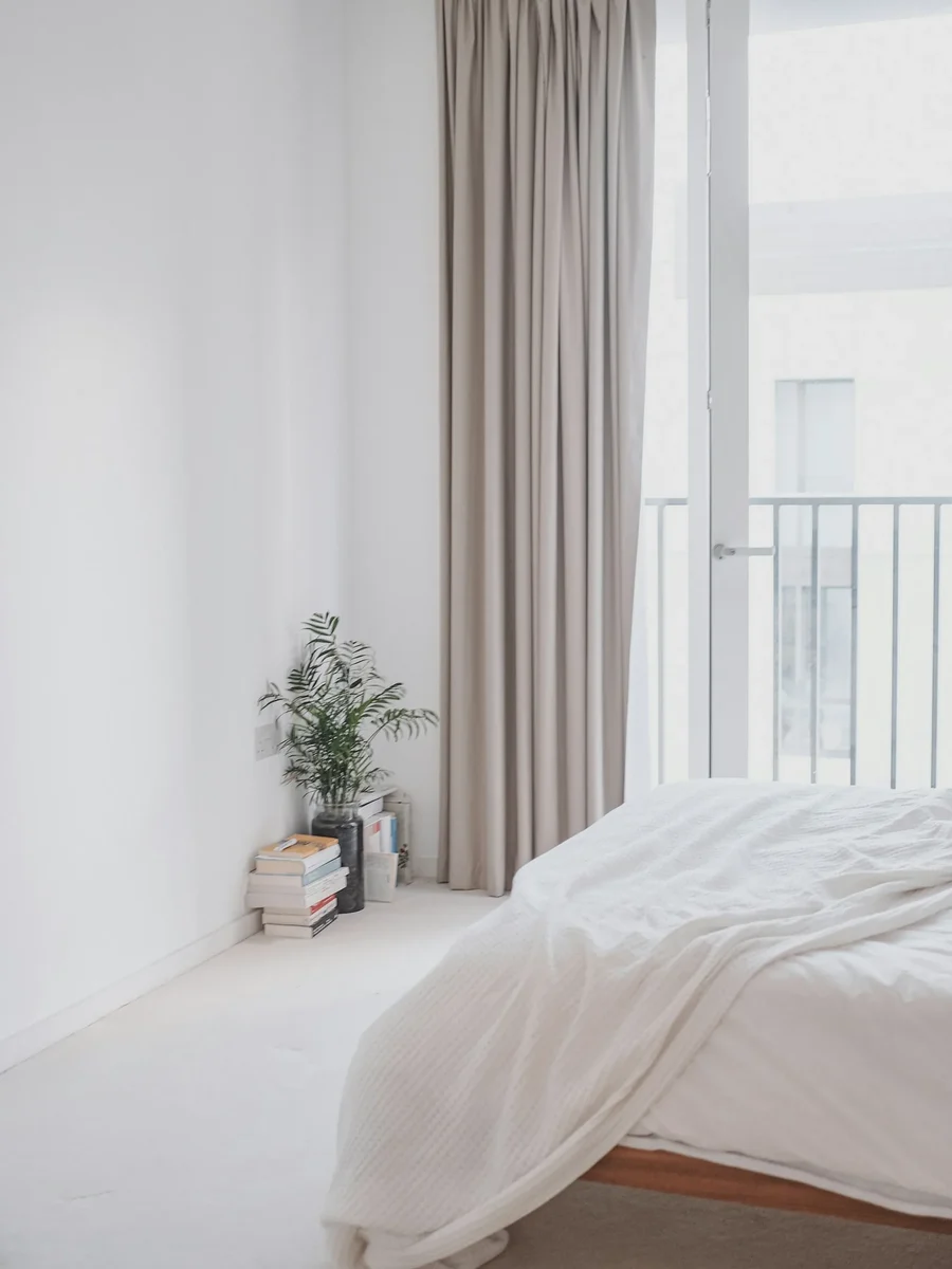 A bright white bedroom with floor-length taupe curtains at a balcony door and a softly made bed