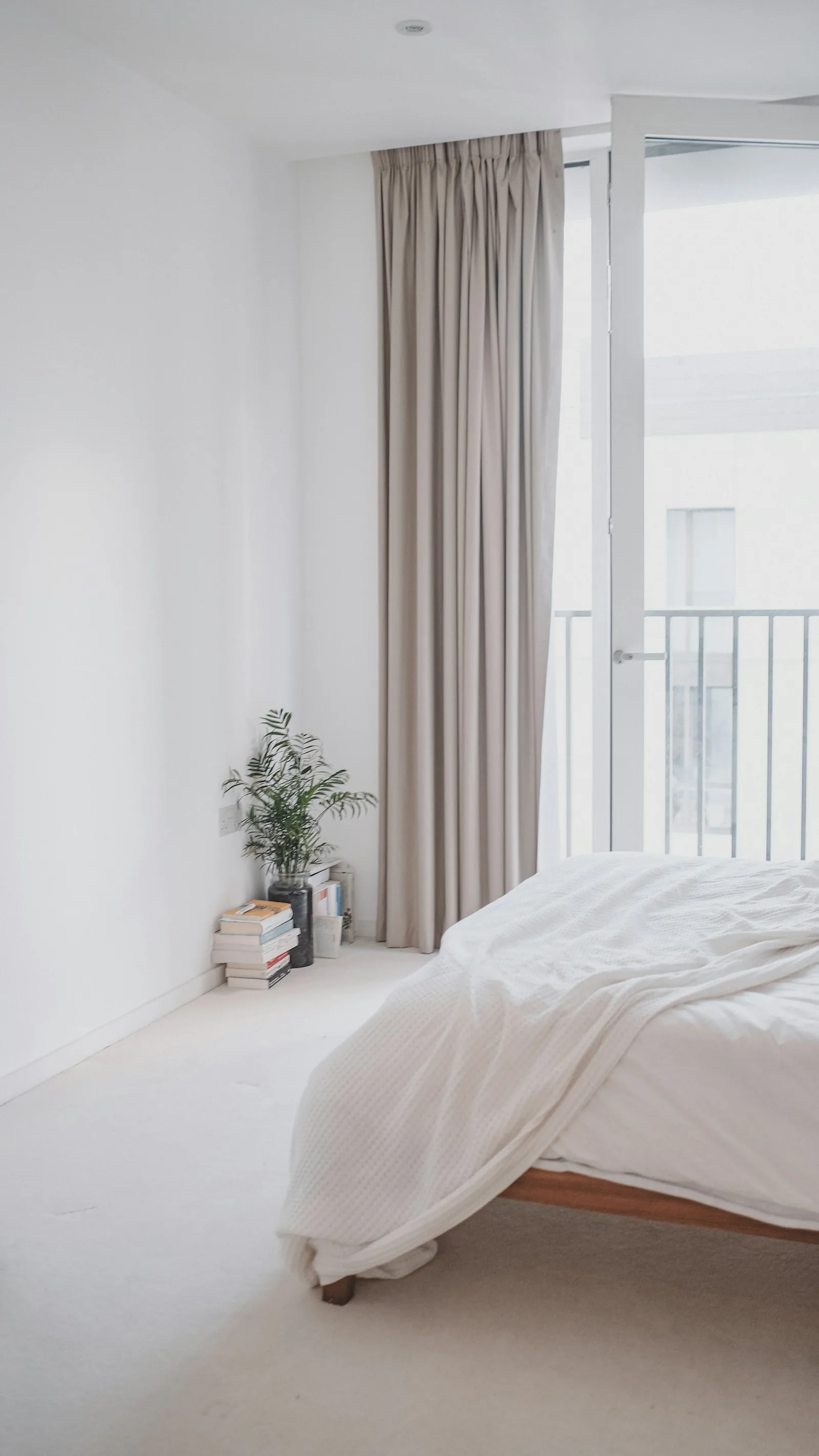 A bright white bedroom with floor-length taupe curtains at a balcony door and a softly made bed