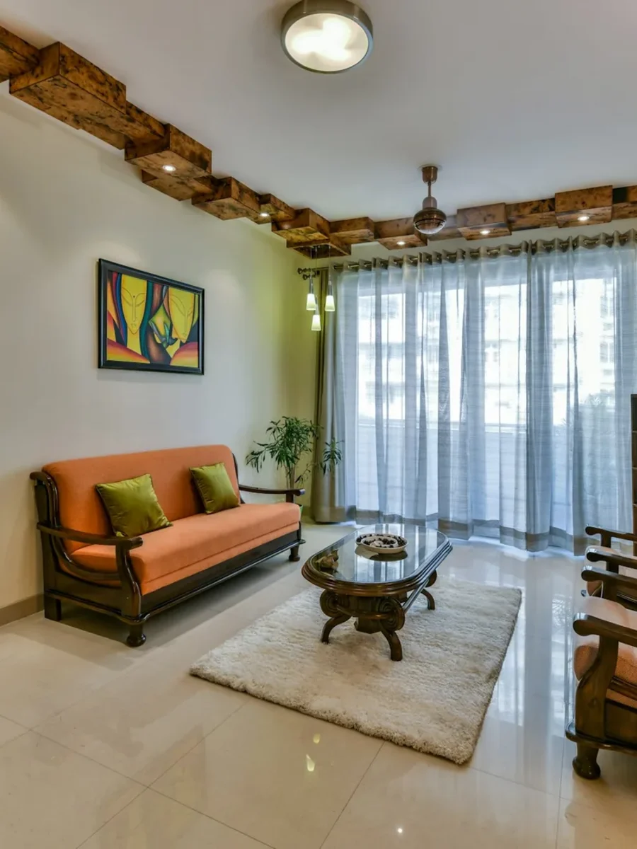 Indian living room with full-height grey sheer curtains, an orange wooden sofa set and a ceiling fan
