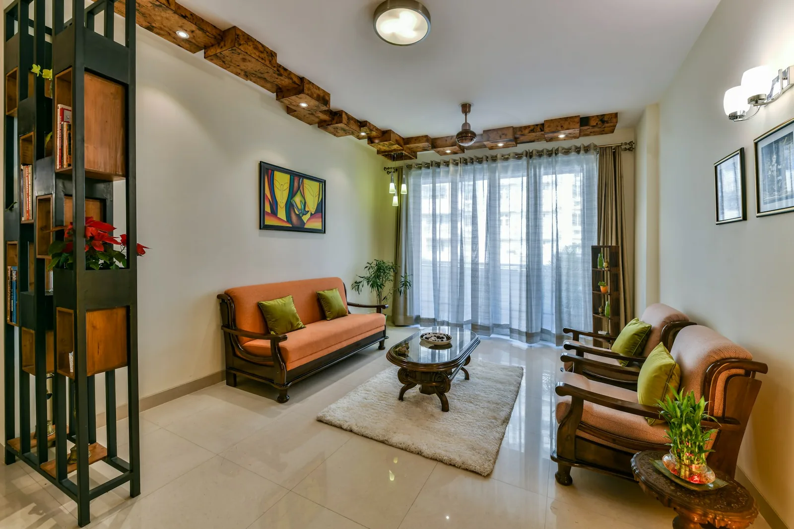 Indian living room with full-height grey sheer curtains, an orange wooden sofa set and a ceiling fan