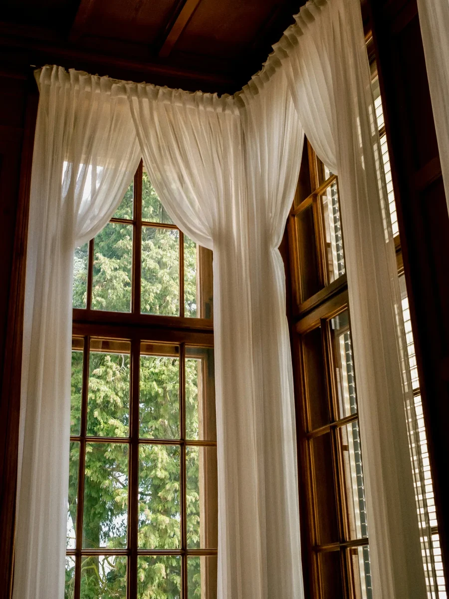 Floor-length white sheer curtains gathered at a tall timber-framed window beneath a dark wooden ceiling