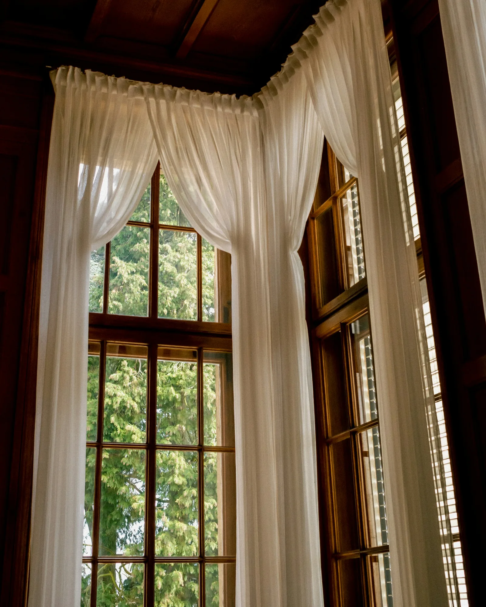 Floor-length white sheer curtains gathered at a tall timber-framed window beneath a dark wooden ceiling