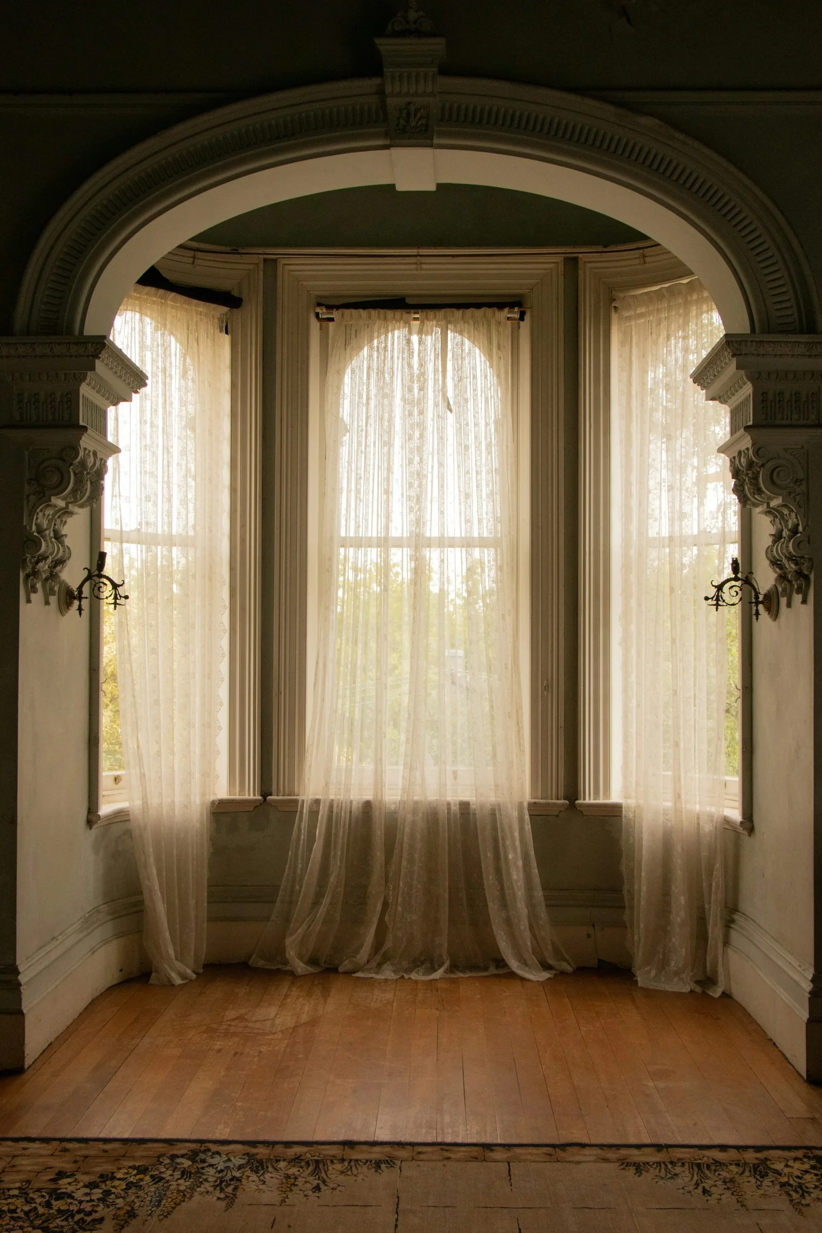 Lace sheers hanging in a tall arched bay window of an old mansion with timber floor and plaster mouldings