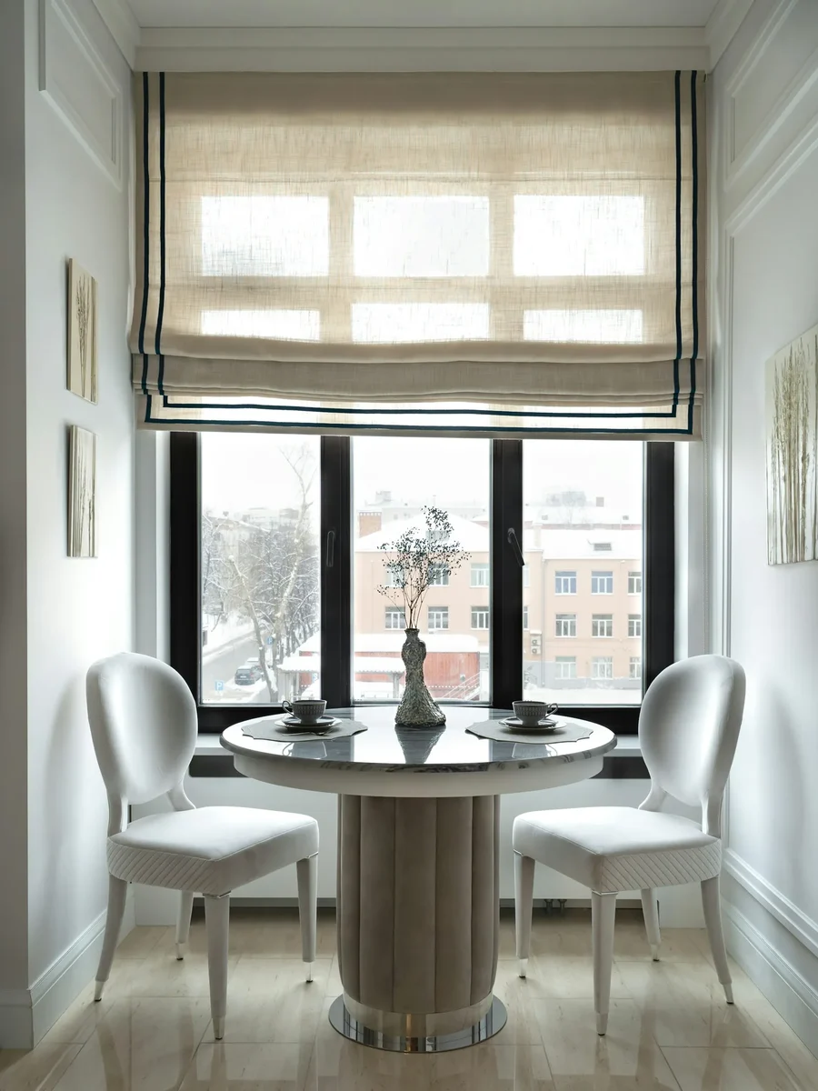 A linen Roman blind with dark trim above a window, over a small round table and two white chairs