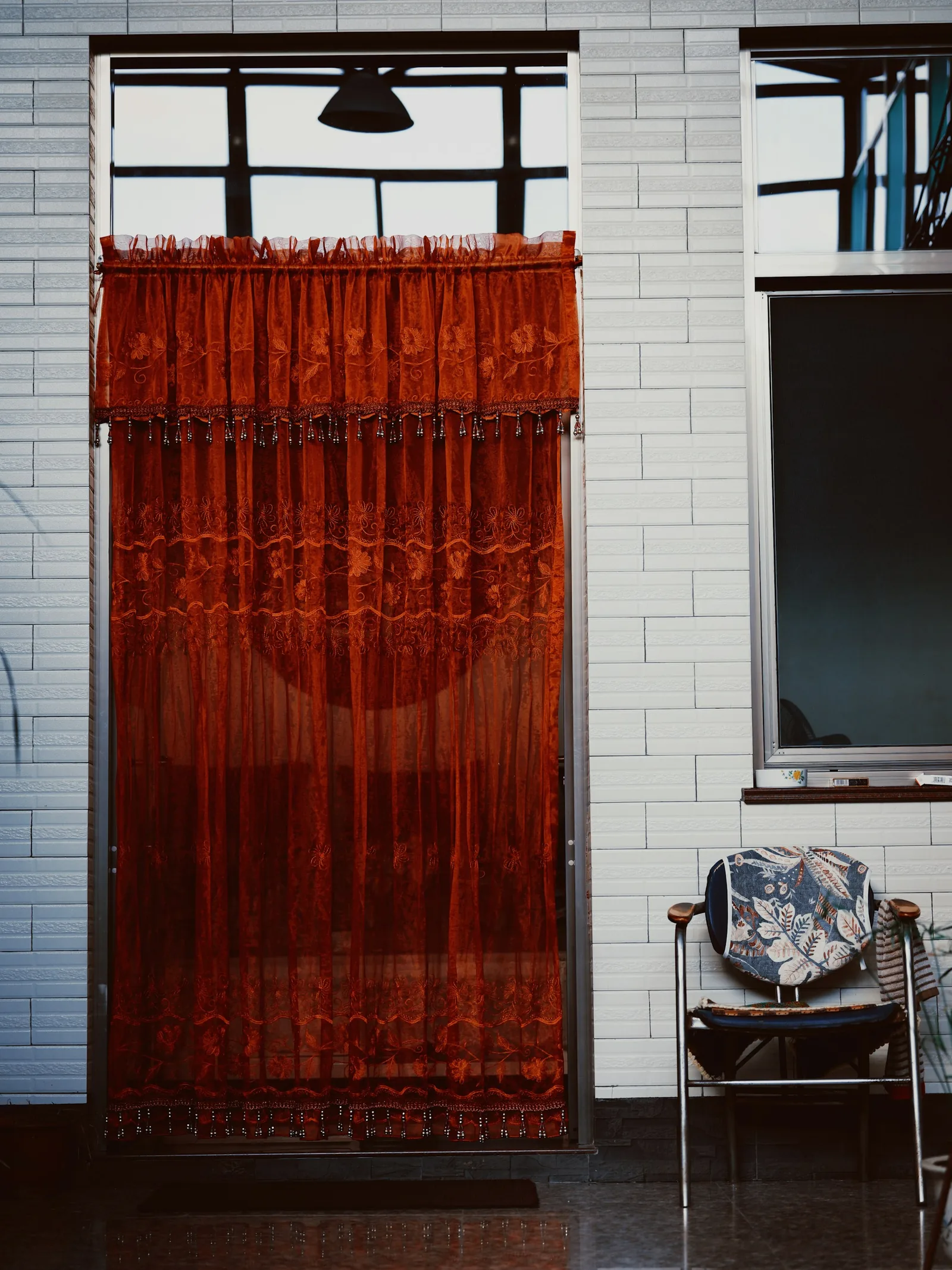 A rust-red patterned curtain hung across a full-height doorway beside a white tiled wall and a chair