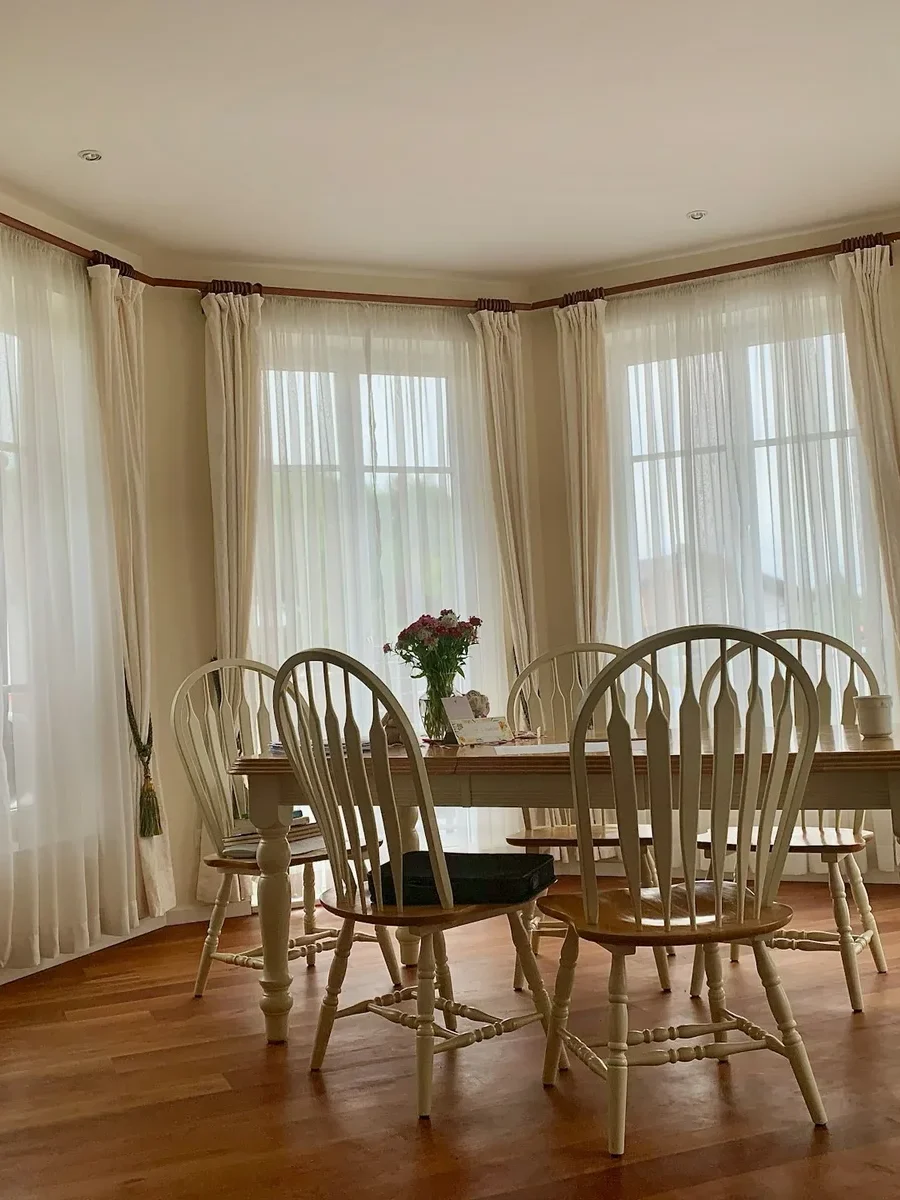 A bay-window dining room with white sheers behind tied-back cream drapes around a wooden table
