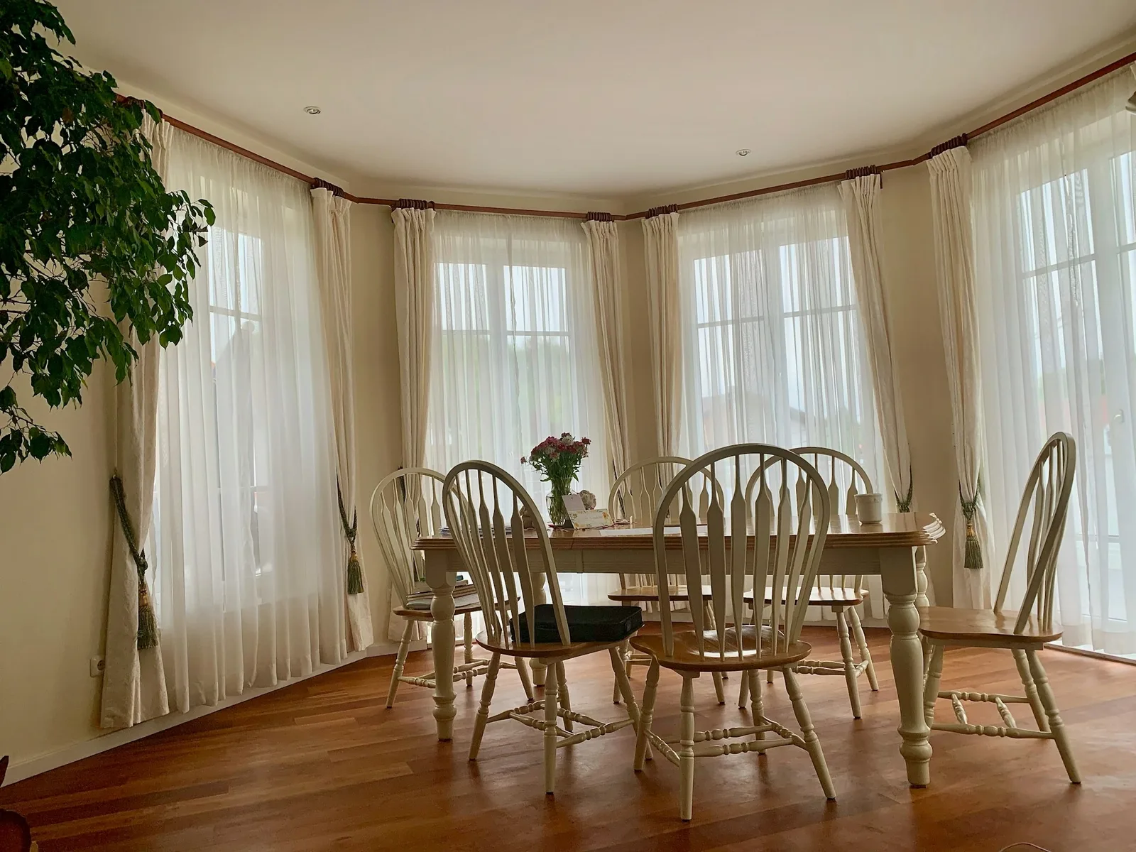 A bay-window dining room with white sheers behind tied-back cream drapes around a wooden table