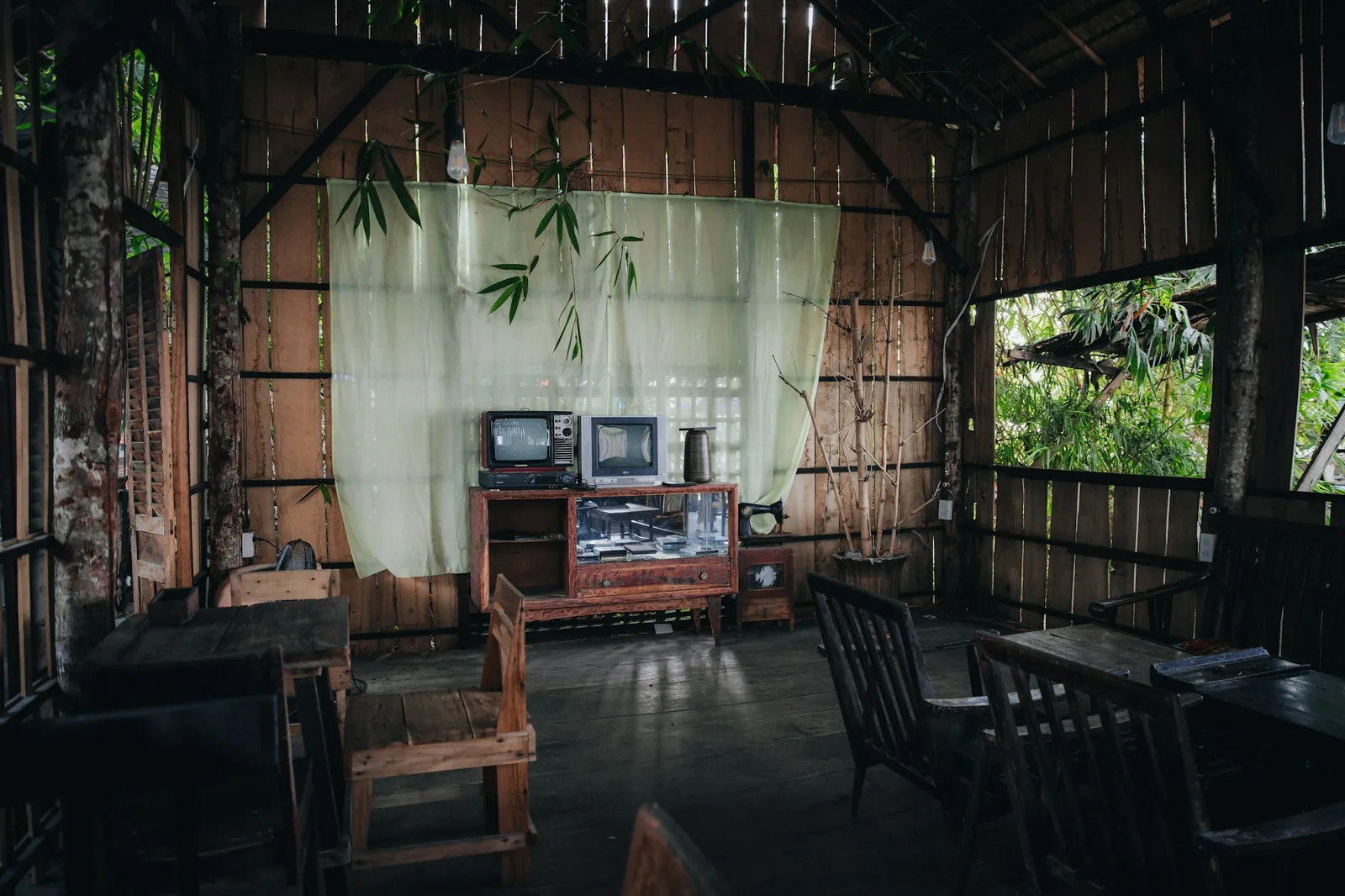 A rustic open-sided timber house with sheer curtains, wooden furniture and greenery visible through the walls