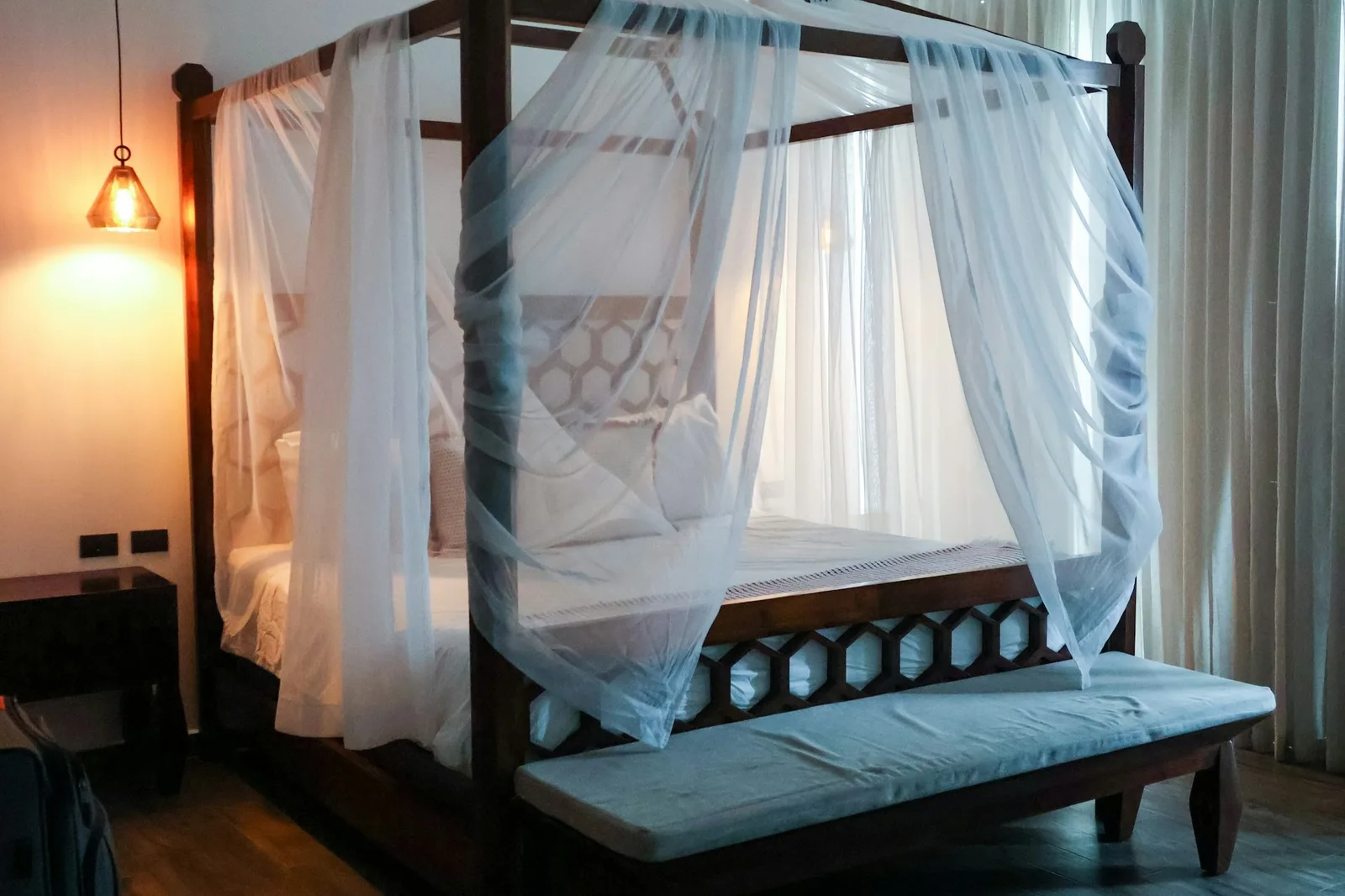 A dark wooden four-poster bed draped with sheer white netting, with a cushioned bench at its foot