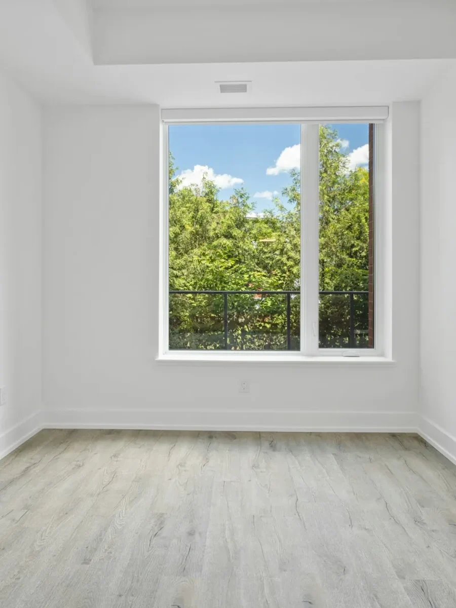 Empty unfurnished room with white walls, pale wood-look floor and a single window onto trees