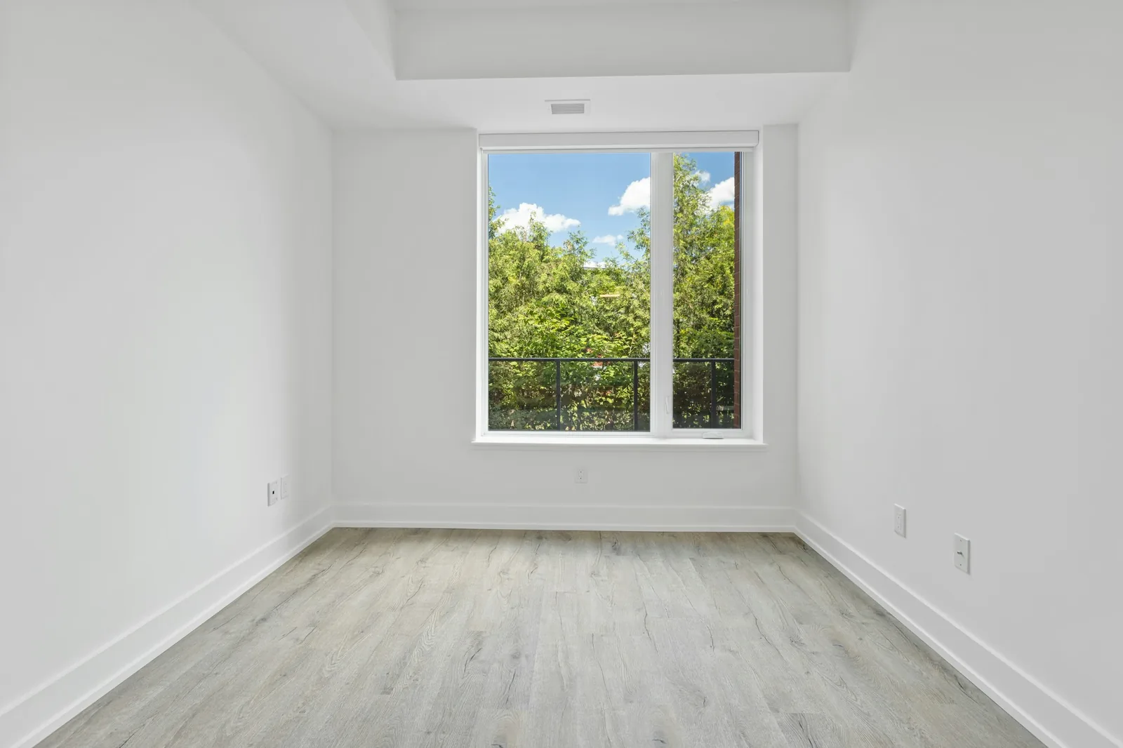 Empty unfurnished room with white walls, pale wood-look floor and a single window onto trees