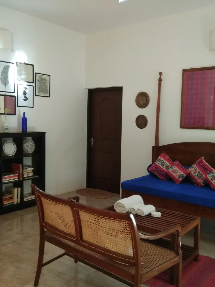 A Goan sitting room with a cane-backed teak bench, a carved daybed with blue mattress and patterned cushions