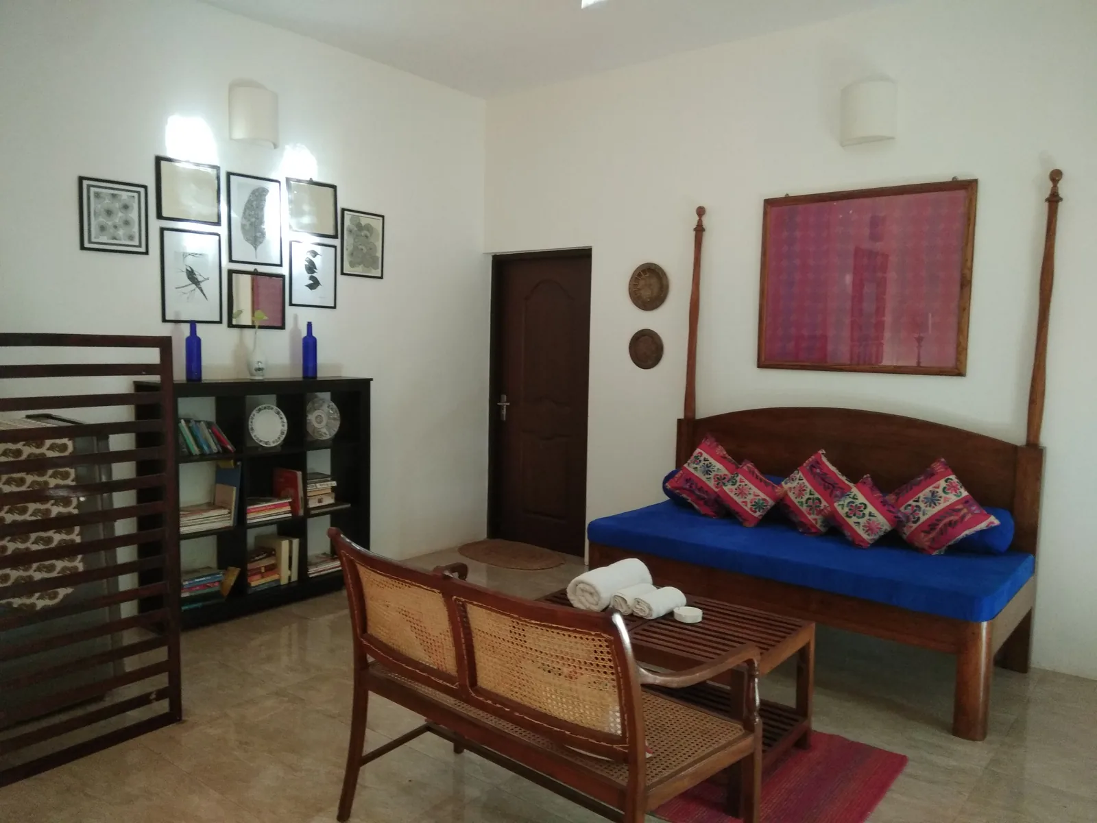 A Goan sitting room with a cane-backed teak bench, a carved daybed with blue mattress and patterned cushions