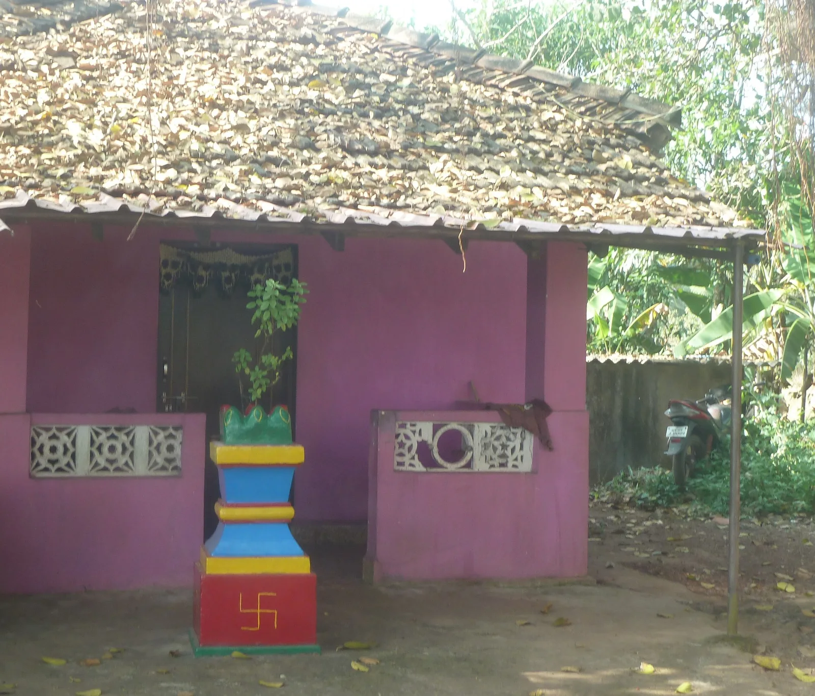 A pink-walled Goan Hindu home with a tiled verandah roof and a painted tulsi vrindavan in the forecourt