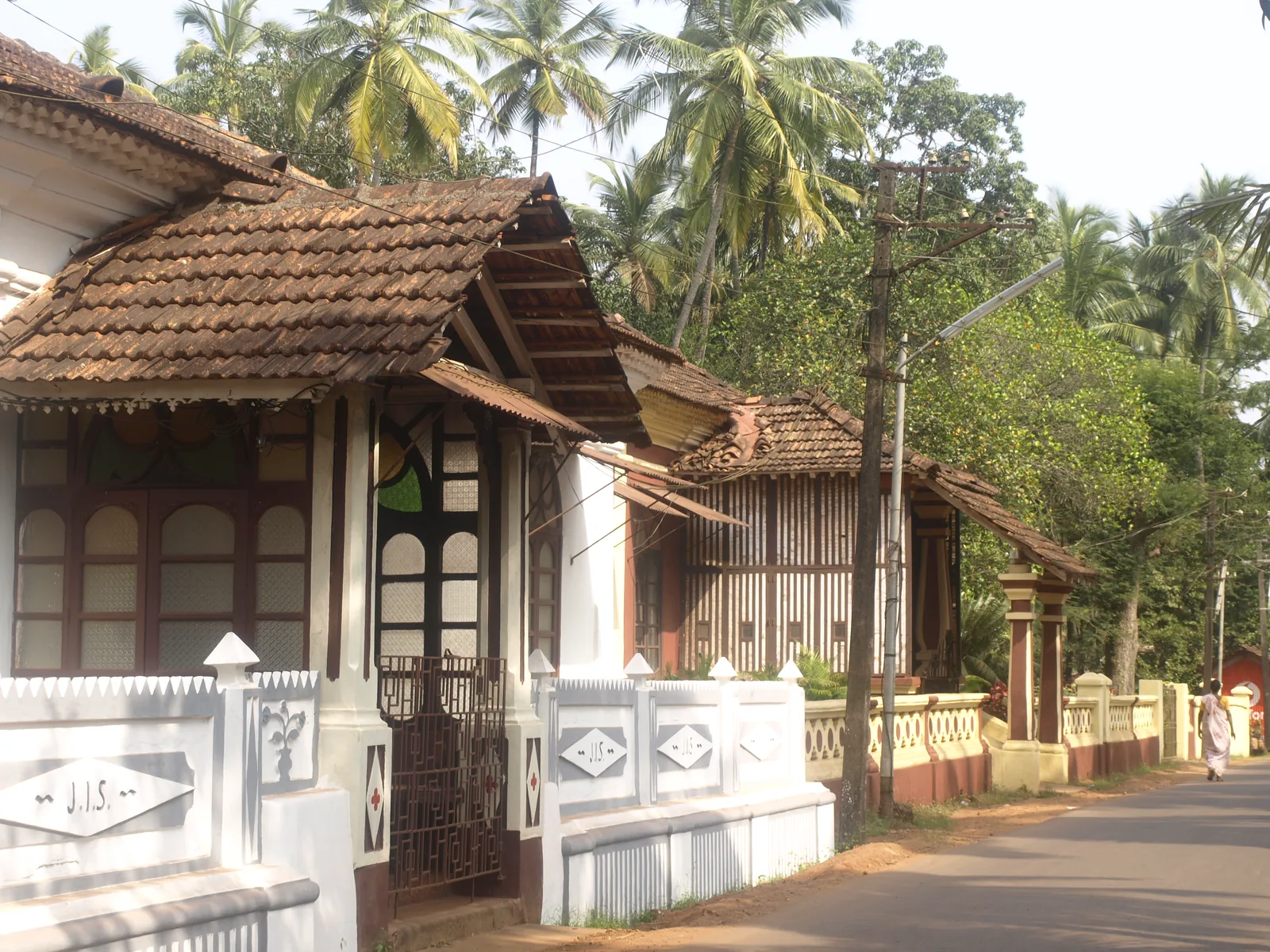 Tiled-roof village houses behind white compound walls on a palm-lined road in Saligao, Bardez