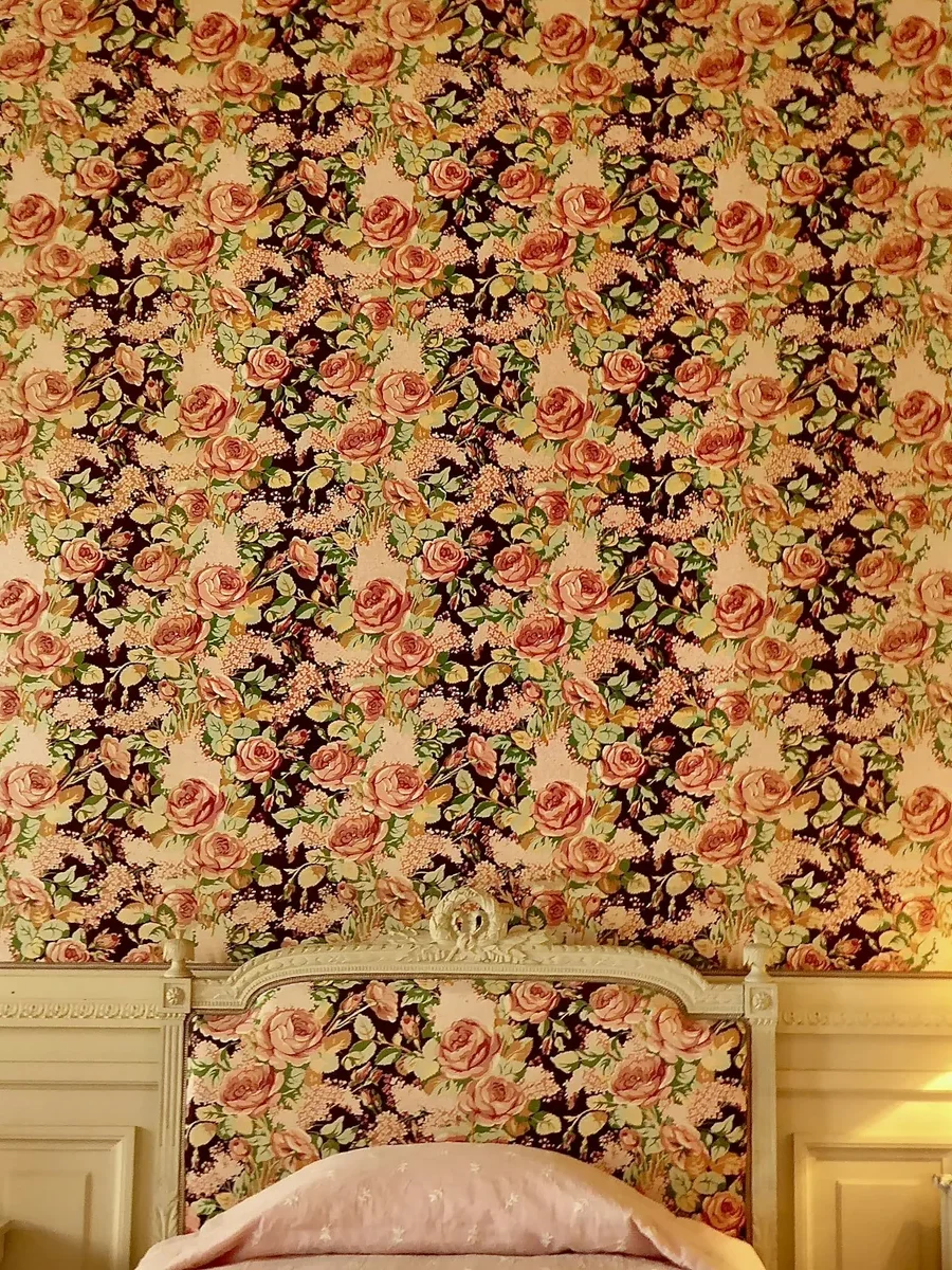 A bed with an upholstered floral headboard matching rose-patterned wallpaper, lit by a bedside lamp