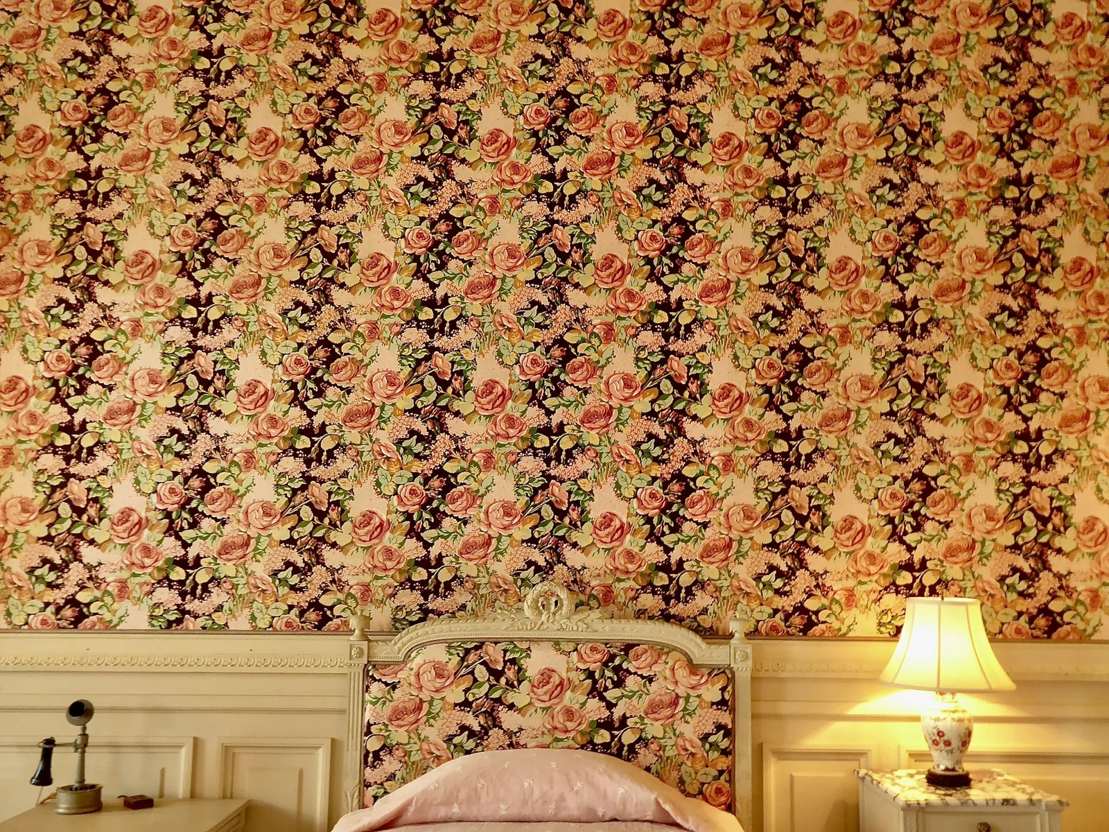 A bed with an upholstered floral headboard matching rose-patterned wallpaper, lit by a bedside lamp