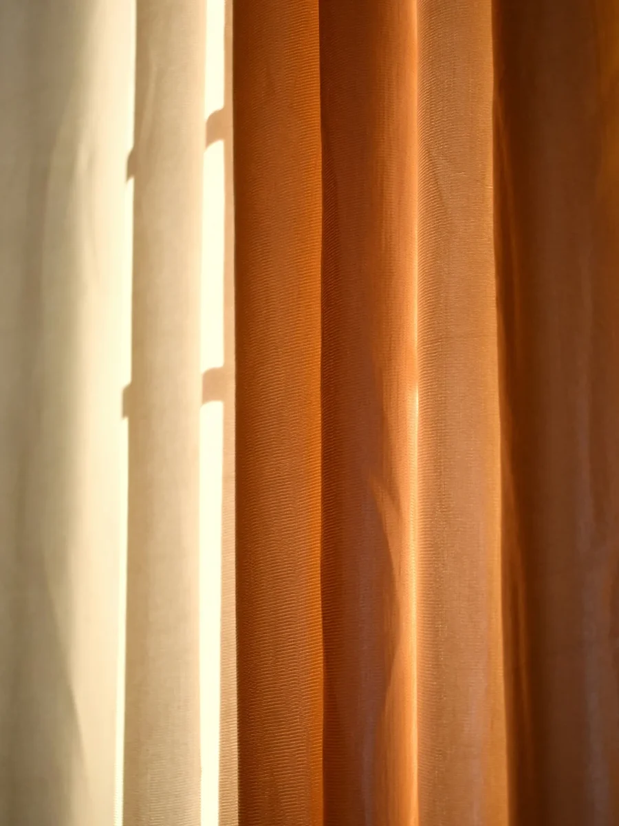 Afternoon sun striking a rust-coloured curtain beside a pale lined panel, showing light and heat on fabric