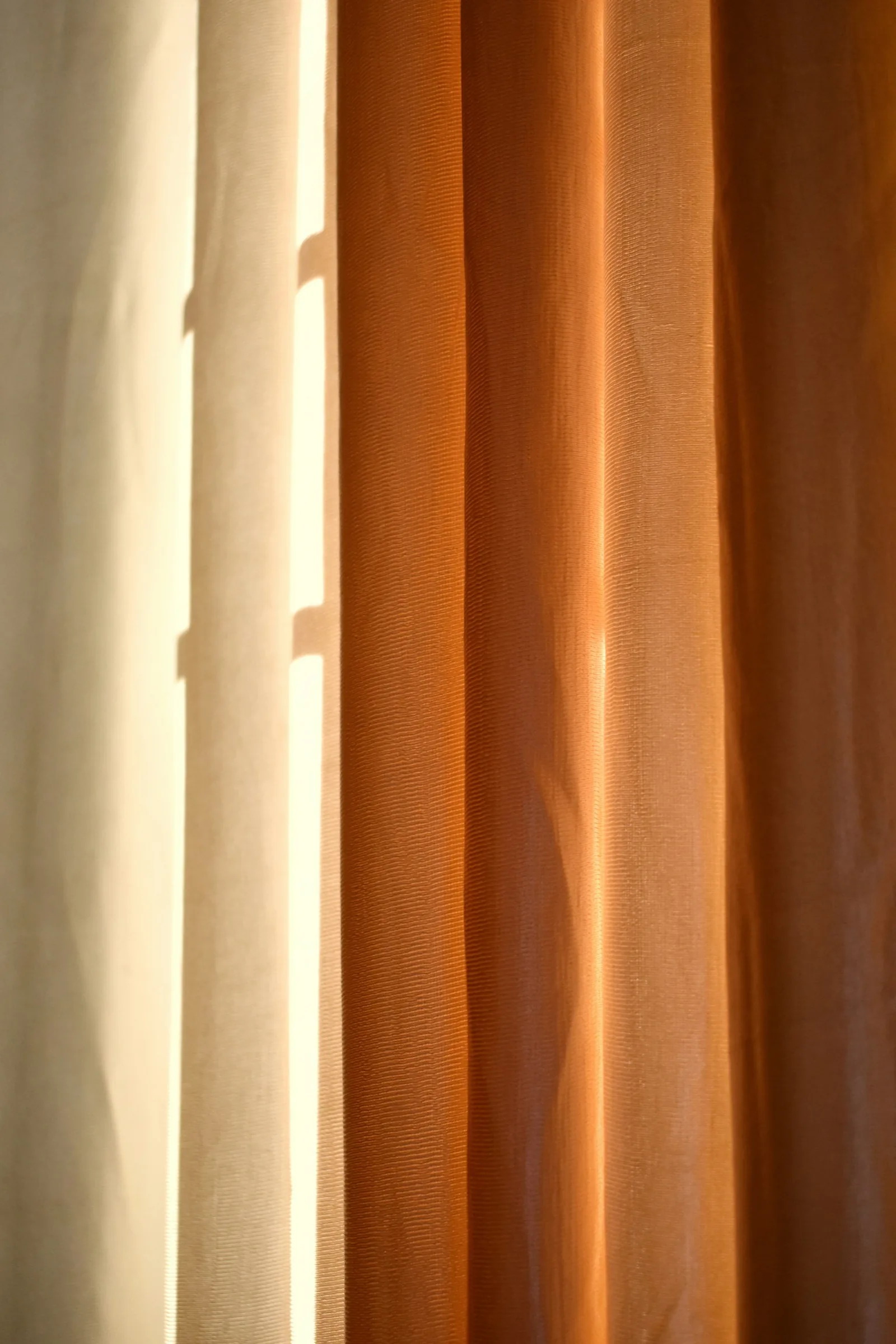 Afternoon sun striking a rust-coloured curtain beside a pale lined panel, showing light and heat on fabric