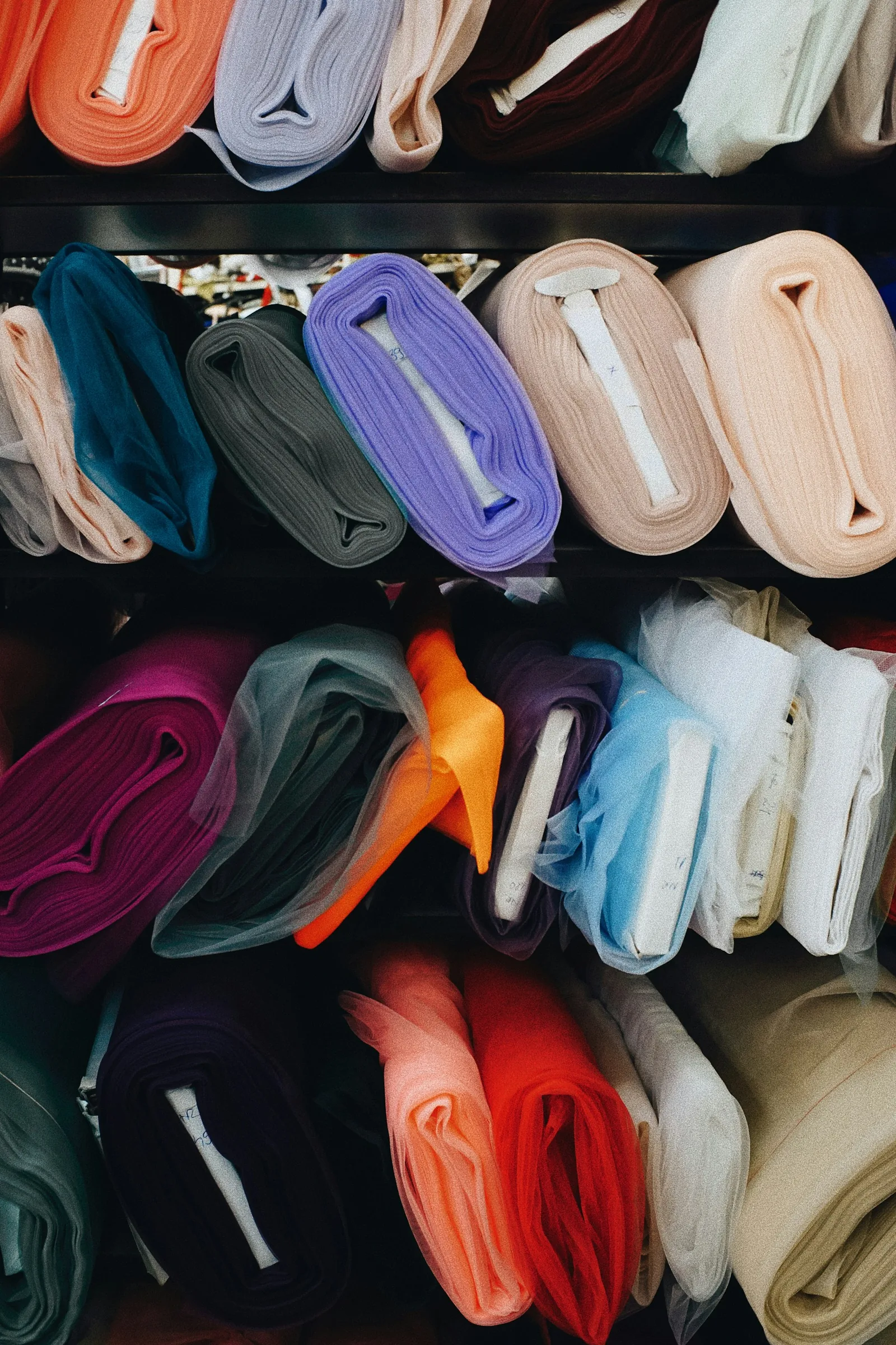 Rolls of fabric in many colours, from sheer tulle to solid weaves, stacked on shop shelves